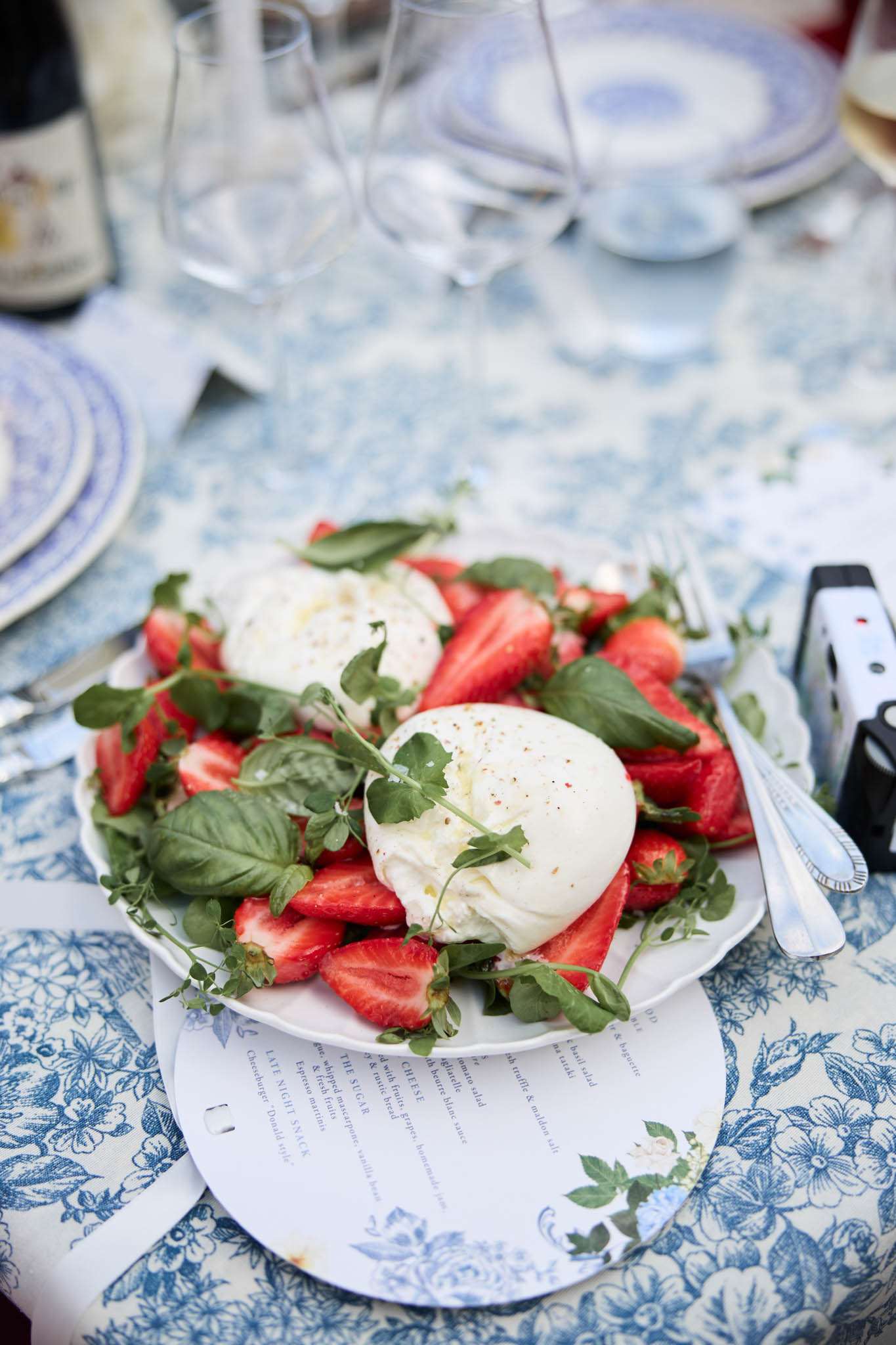 A close-up detail shot of a plated starter course at a wedding reception dinner, showing a burrata and strawberry salad garnished with fresh basil and herb leaves, drizzled with olive oil and cracked pepper, served on a white plate. The plate rests on a printed menu card and is set on a blue and white floral toile tablecloth, with blue and white patterned charger plates, silver cutlery, crystal wine glasses, and a dark wine bottle visible in the background. The table styling follows a classic blue and white colour palette with a French country aesthetic. The shot is taken at a slight angle, focused on the food with the surrounding table setting softly blurred.