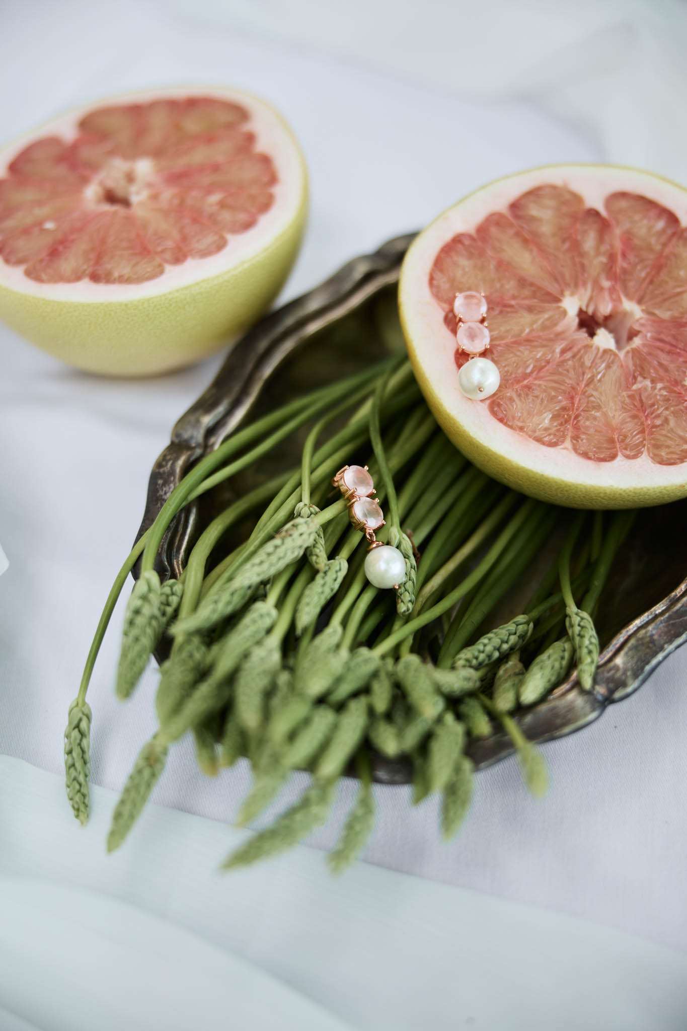 Rose gold drop earrings with pink gemstones and pearl drops on silver tray with grapefruit half and wheat stems