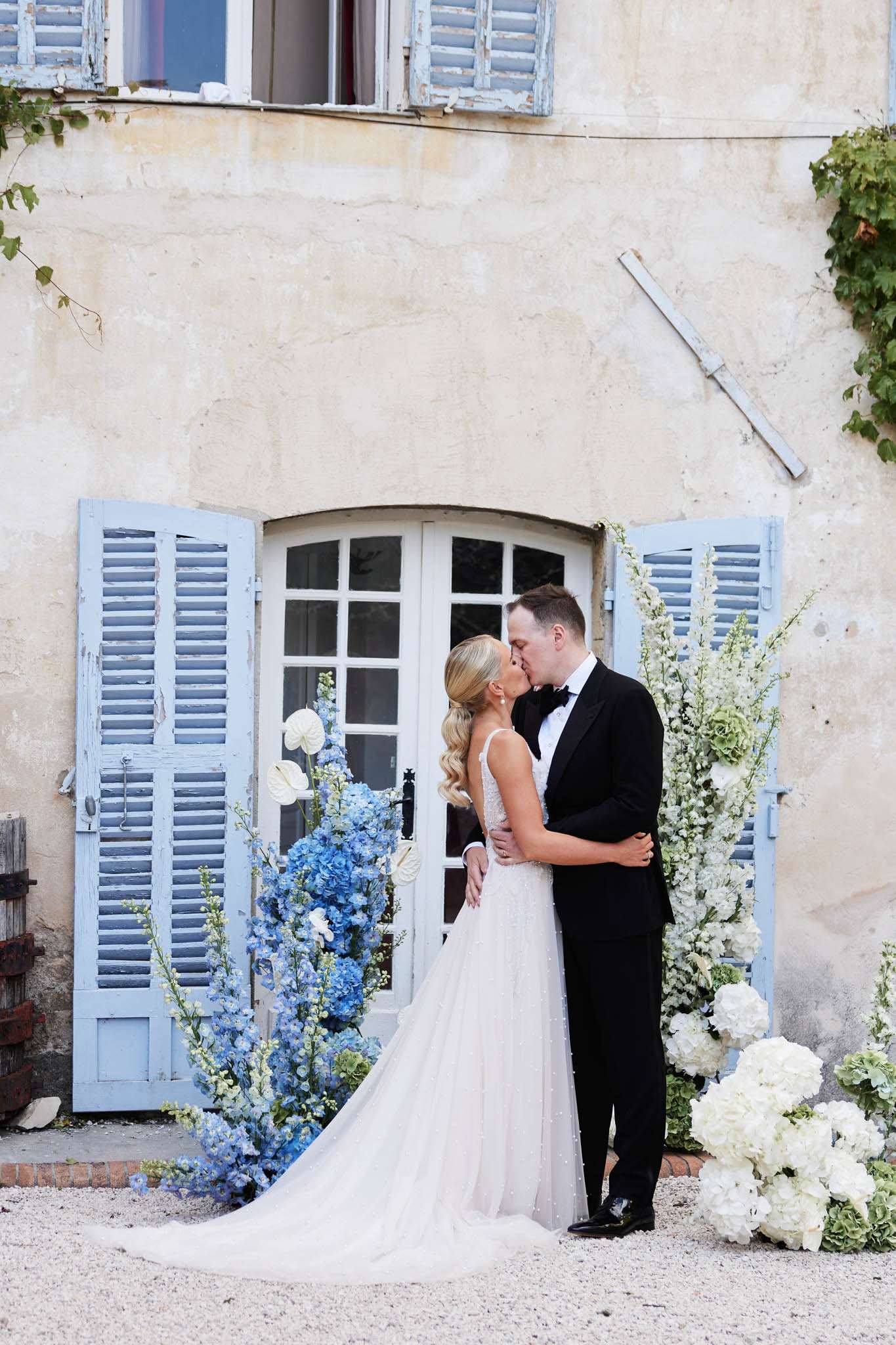 A couple shares a kiss in an outdoor portrait in front of a French country house facade featuring aged render and powder blue painted louvred shutters flanking an arched white door. The bride wears a fitted, beaded ivory gown with a V-neckline and a long train, while the groom is dressed in a classic black tuxedo with a bow tie. Flanking them are tall floral arrangements featuring blue hydrangeas and blue delphiniums on the left, and white delphiniums and white hydrangeas on the right, with additional clusters of white and sage green hydrangea blooms placed on the gravel ground. The styling palette is blue, white, and soft green, giving a classic French garden feel, and the shot is a full-length portrait taken from a slight distance.