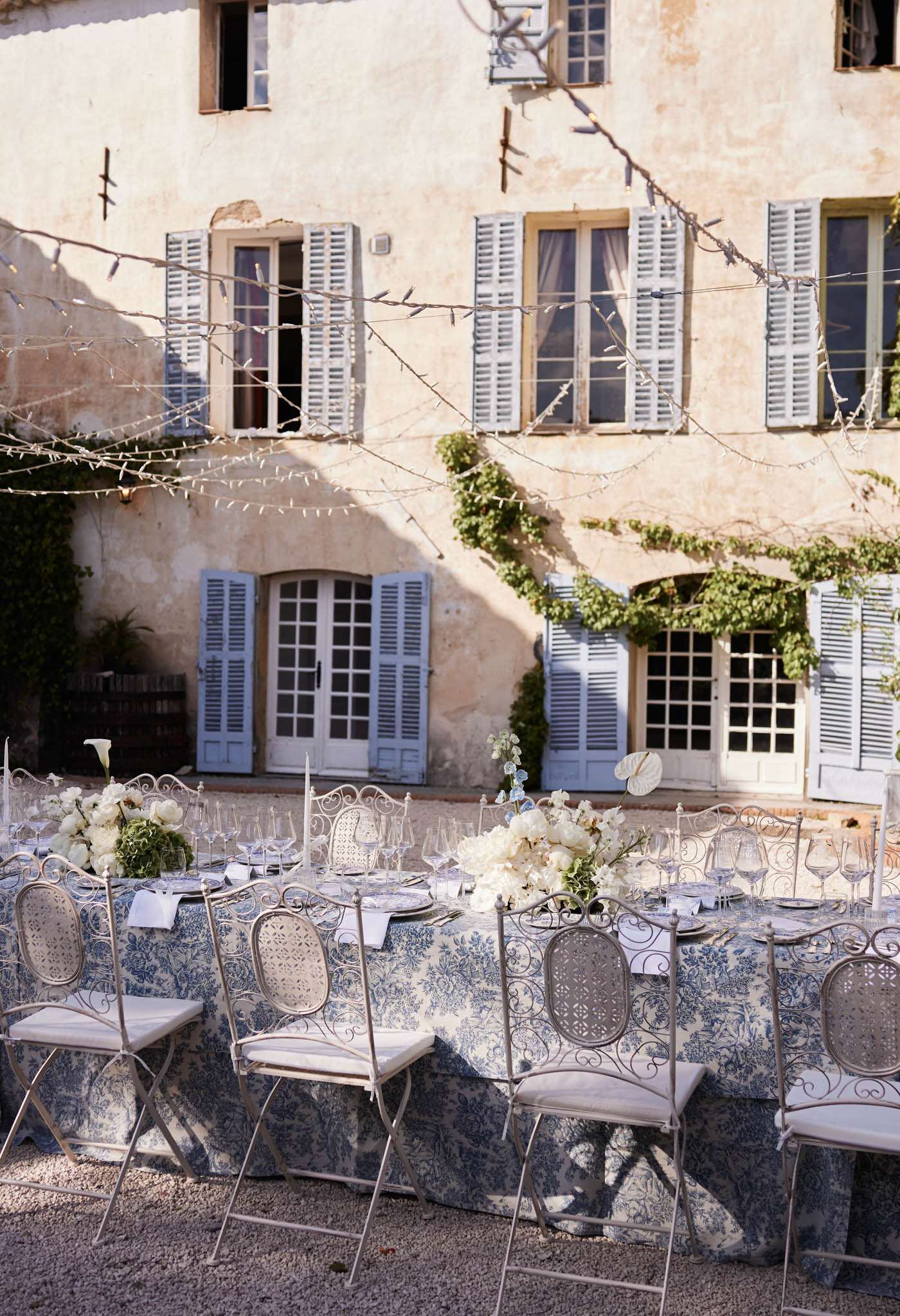 An outdoor wedding reception table is set in the courtyard of a French mas or bastide-style property, featuring pale ochre rendered walls and grey-blue painted shutters across multiple windows and French doors. The long rectangular dining table is dressed in a blue-and-white toile de Jouy printed tablecloth, surrounded by ornate grey wrought-iron bistro-style chairs with cane backs and cream seat cushions. Centerpieces consist of low, lush arrangements of ivory peonies, white ranunculus, and white calla lilies with green foliage, placed at intervals along the table alongside tall taper candles and clusters of crystal wine glasses and place settings. Fairy lights are strung overhead between bare-branched trees crossing in front of the building facade. The overall styling palette is blue, white, and grey with a classic Provençal aesthetic. Wide shot capturing both the full table setting and the building facade as backdrop. Potential venue feature image.