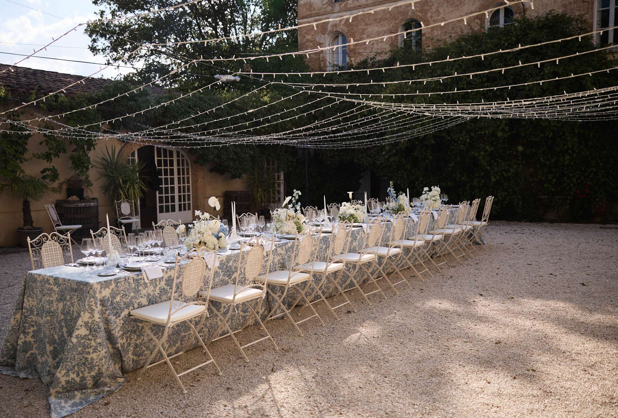 Toile de Jouy table with blue hydrangeas white roses silver candlesticks wrought-iron rattan chairs under fairy light canopy