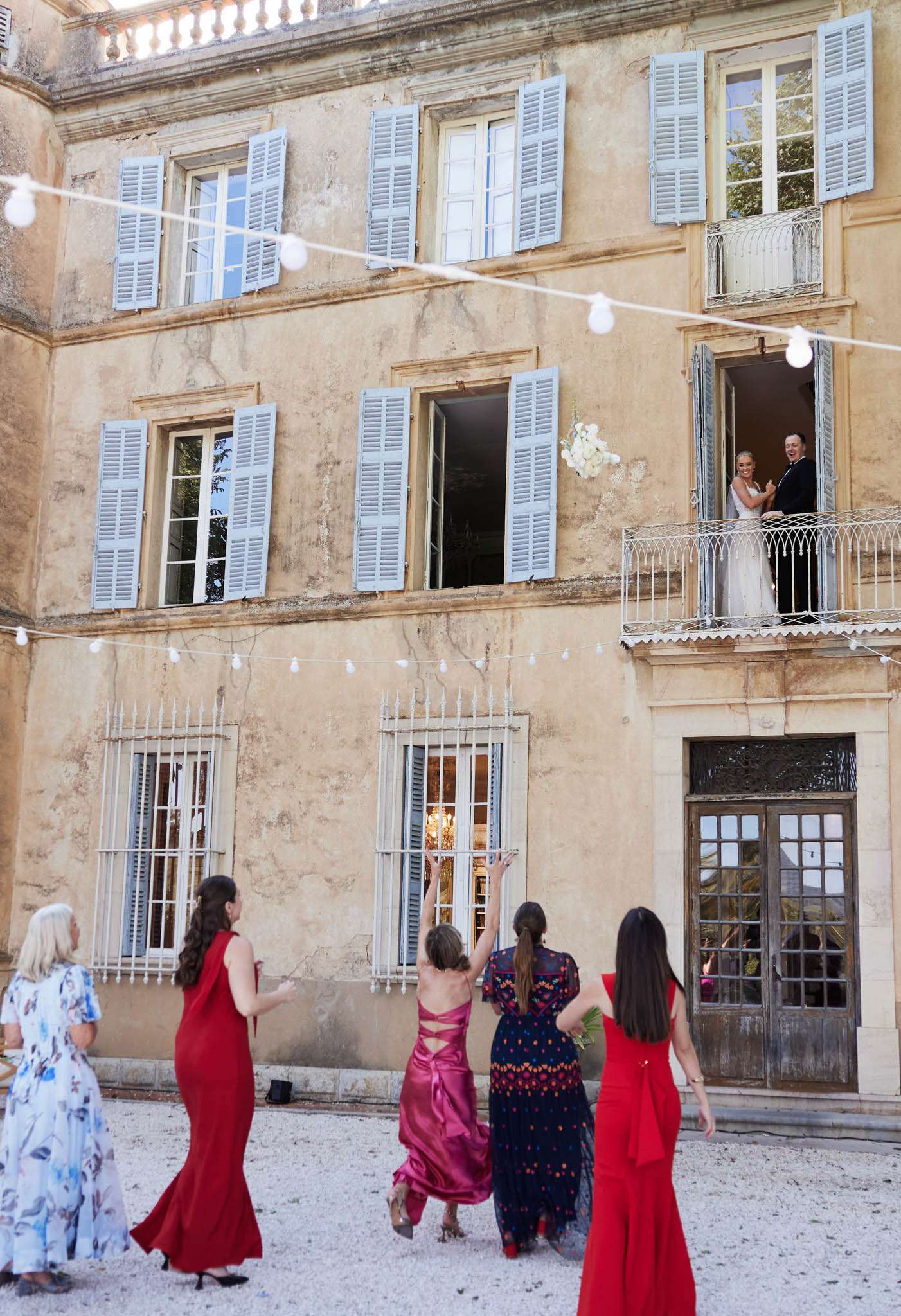 Bouquet toss from juliet balcony with five guests in red and fuchsia gowns reaching below string lights
