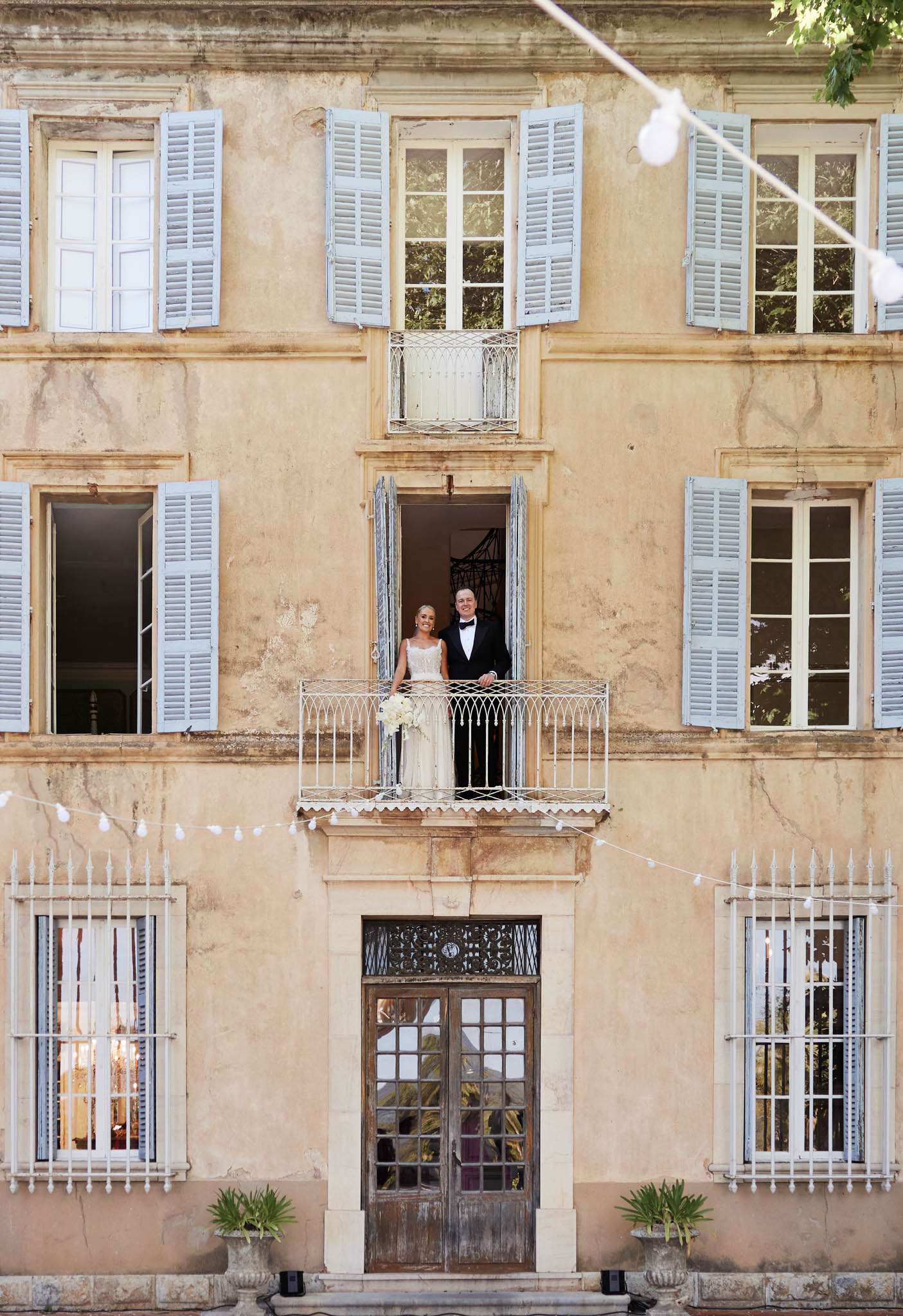 Couple poses on wrought-iron balcony of ochre chateau with blue shutters and globe lights at dusk