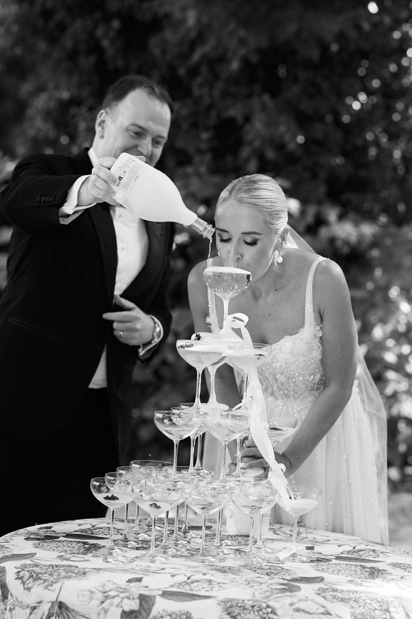 Black and white photo of couple pouring champagne tower, groom in dinner jacket, bride in beaded gown