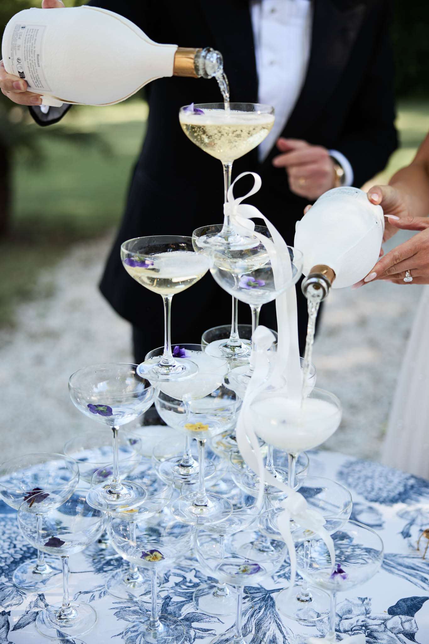 Champagne tower of coupe glasses garnished with edible flowers being filled from two magnum bottles