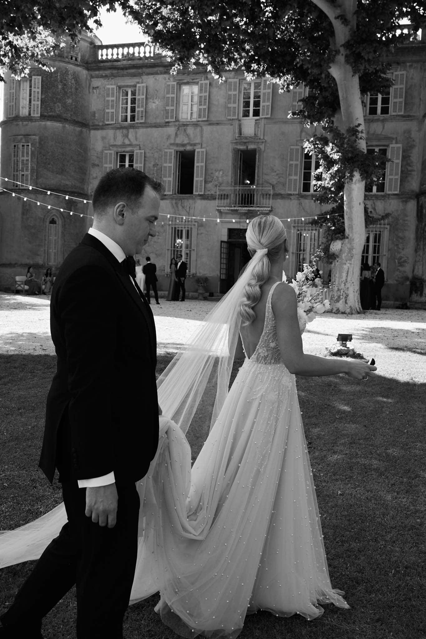 A black-and-white outdoor portrait of a bride and groom walking across the grounds of a large French château, photographed from behind and to the side. The groom wears a dark tuxedo with a bow tie, while the bride wears a full A-line gown with an allover pearl or bead embellishment detail, a low open back, and a long cathedral-length veil; her blonde hair is styled in a low ponytail with loose waves. She carries a bouquet of light-toned flowers. The château façade behind them is a multi-storey classical French manor with shuttered windows, a balustrade roofline, and string bistro lights strung across the courtyard. Several guests and what appears to be a wedding party are visible in the background near the building's entrance. The image is a medium-wide candid shot with strong contrast between the deep dark tones of the groom's suit and the bright white of the bride's gown and veil. Potential venue feature image.