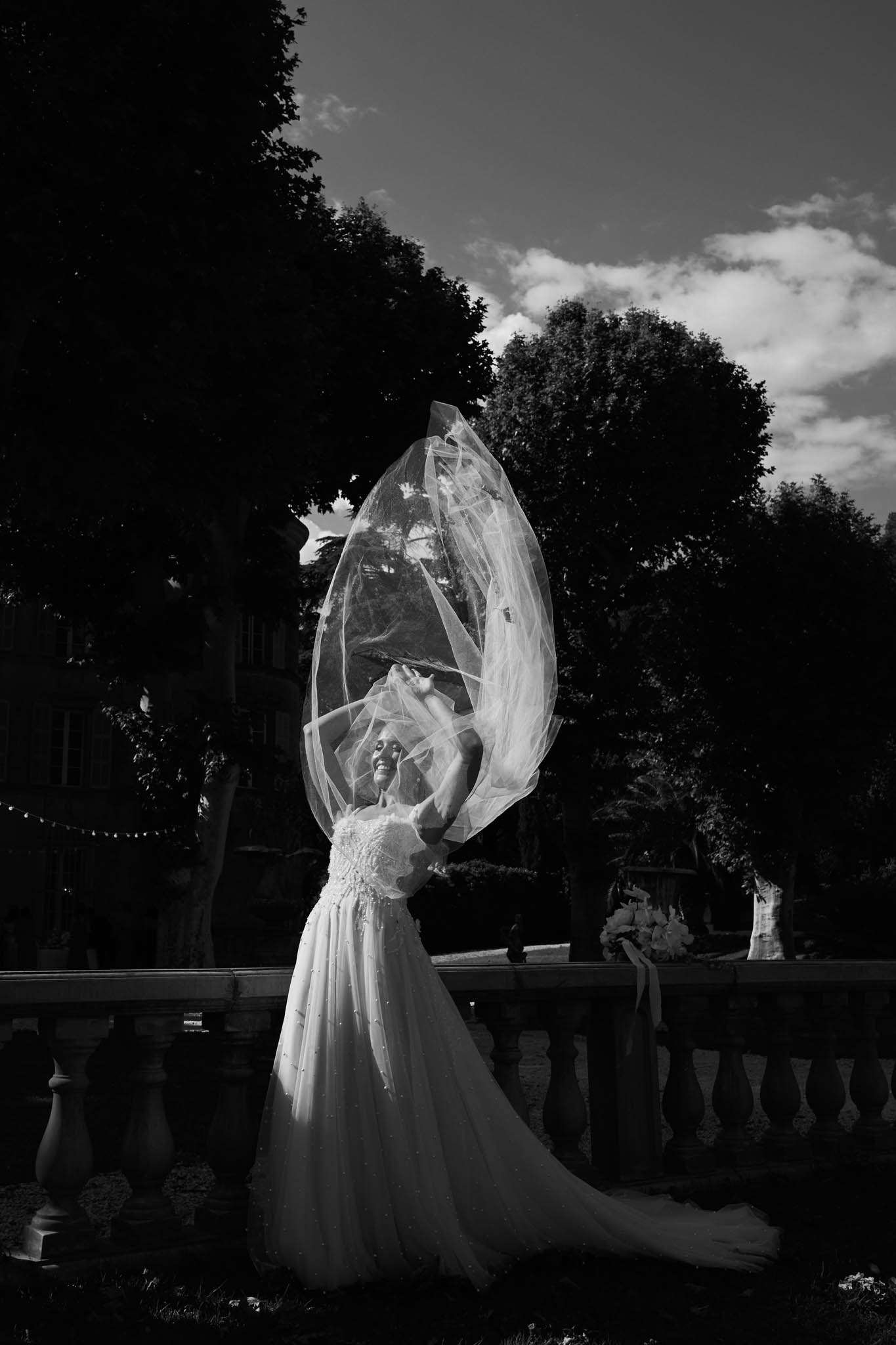 Bride tossing cathedral veil upward beside stone balustrade with string lights in garden in B&W