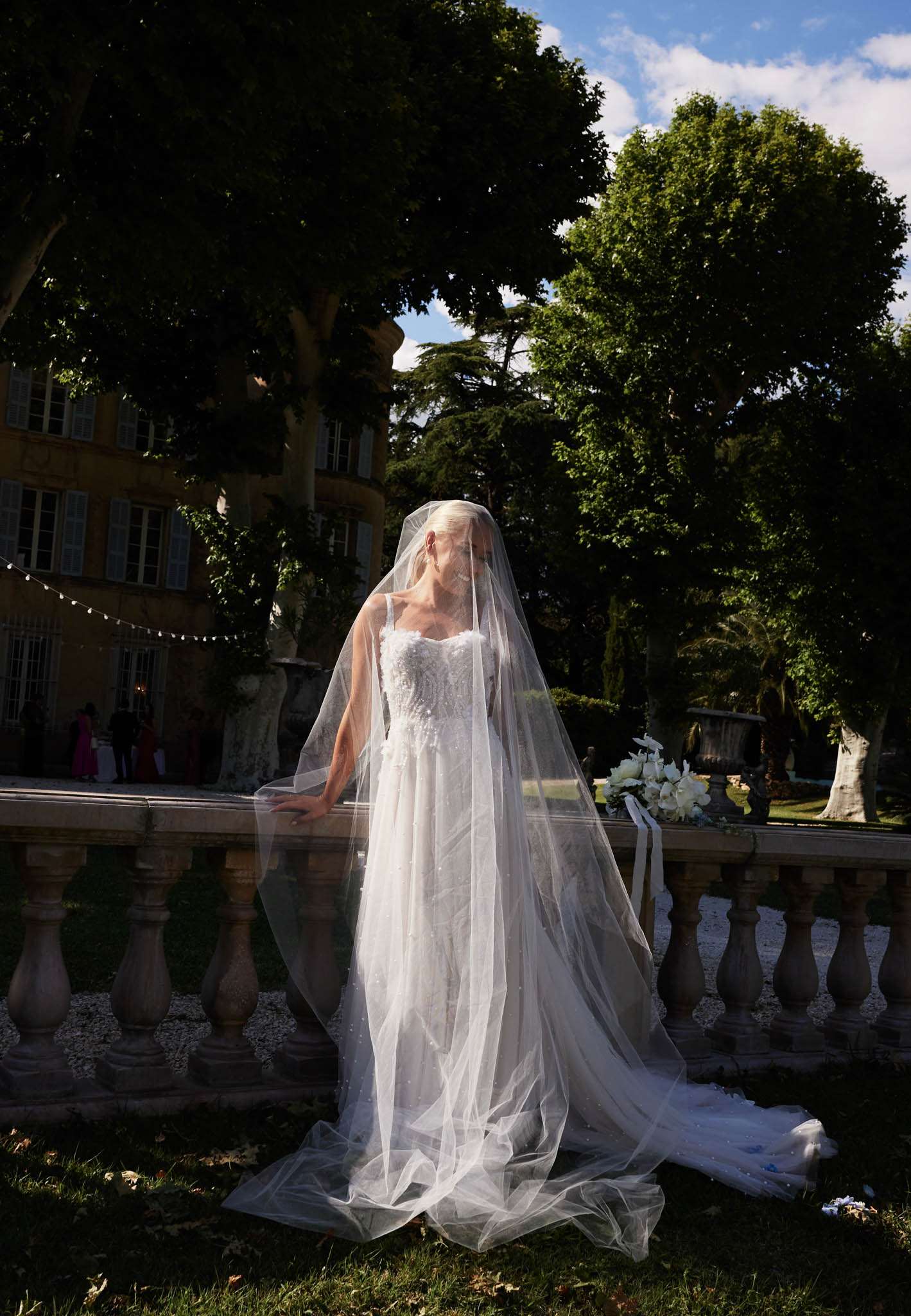 A bridal portrait taken outdoors on a stone balustrade terrace at what appears to be a French château, visible in the background as a yellow ochre-rendered manor house with shuttered windows. The bride stands facing the camera wearing a white A-line gown with a floral appliqué bodice and a long cathedral-length veil dotted with small pearl or crystal accents, which drapes over her face and pools on the ground below. A white bouquet of large blooms, likely white lilies and orchids with trailing white ribbons, rests on the balustrade beside her. In the far background, a small cluster of guests in bright clothing is visible near the château entrance, and a string of fairy lights is strung between trees. The image is a full-length portrait shot with dramatic contrast between deep shadow and bright midday sunlight.