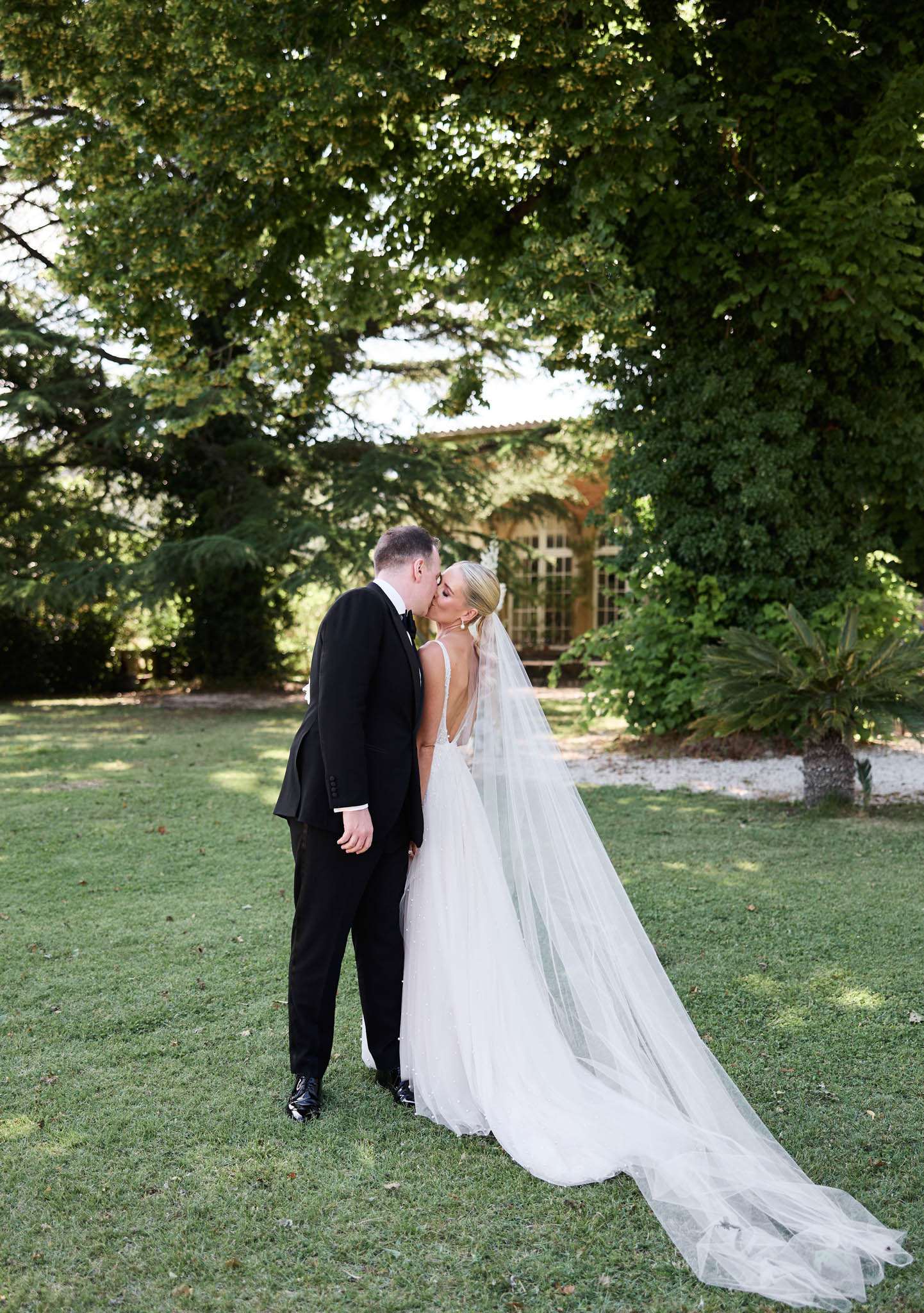 Bride and groom kissing on chateau lawn, bride in cathedral-length veil and groom in black tuxedo, shot from behind
