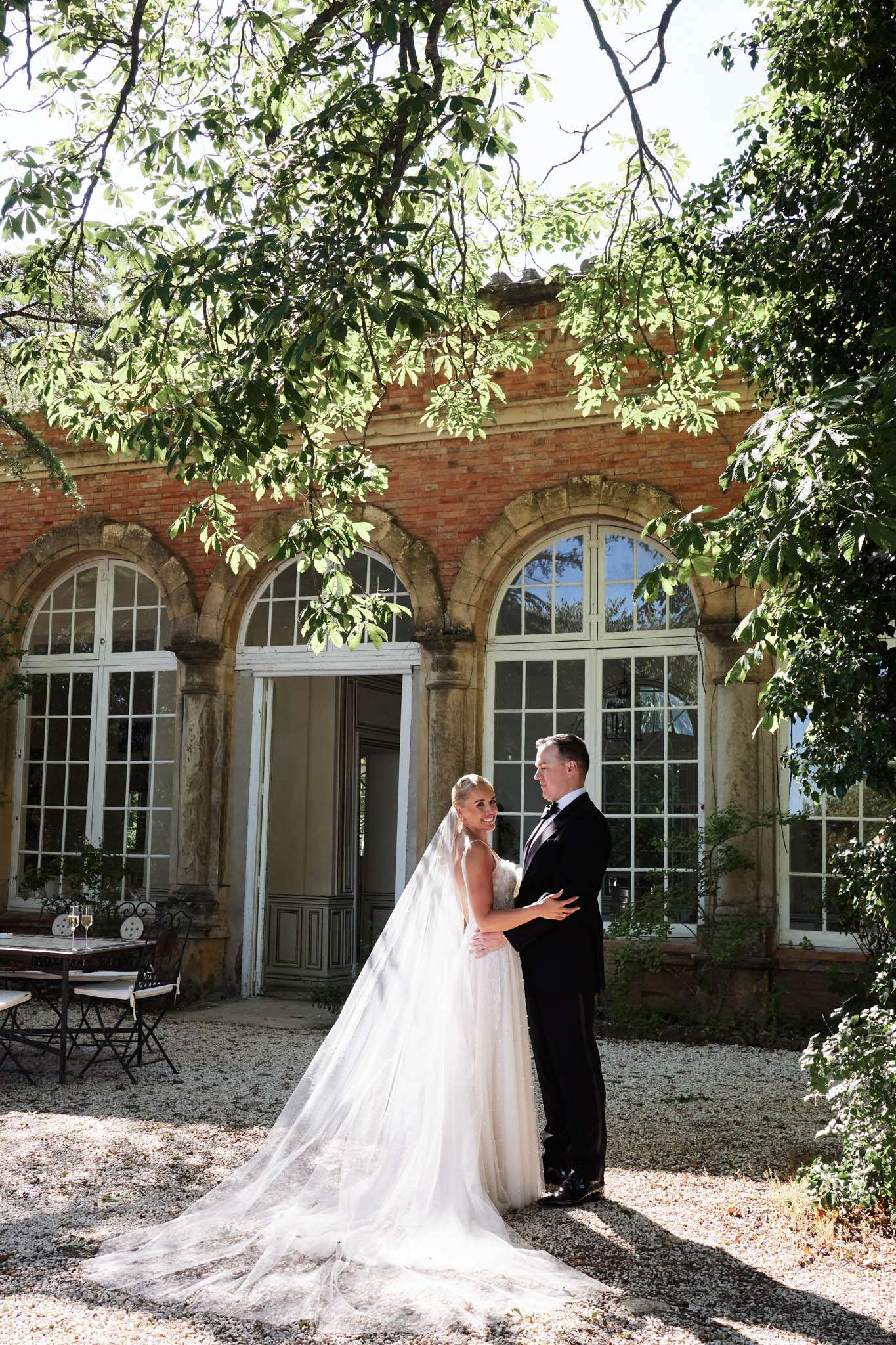 Bride in ivory ballgown with cathedral veil and groom in black tuxedo embracing on chateau gravel courtyard