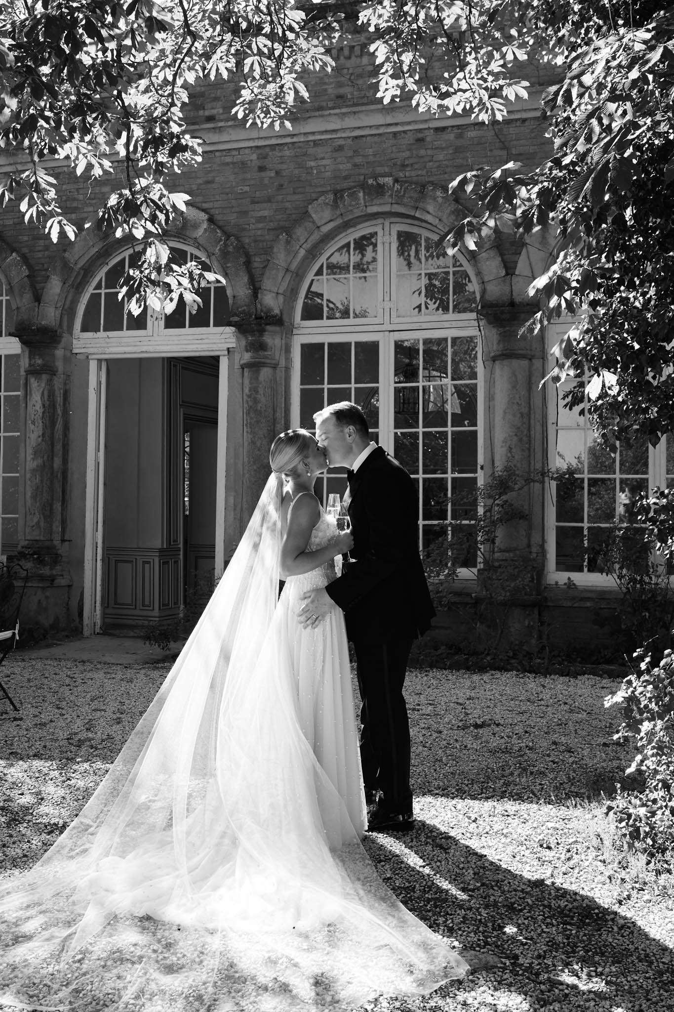 A black-and-white couple portrait taken outdoors in the gravel courtyard of what appears to be a French château or manor house, featuring large arched stone windows and ornate carved surrounds with a set of tall panelled doors to the left. The bride and groom are sharing a kiss; the bride wears a full-skirted ball gown with a fitted bodice and a very long cathedral-length veil that trails across the gravel, while the groom wears a dark suit or tuxedo. Both hold champagne flutes. The image has high contrast with bright highlights on the dress and veil against the deeper tones of the stone facade and foliage. The composition is a mid-to-full-length portrait with tree branches framing the upper portion of the frame.