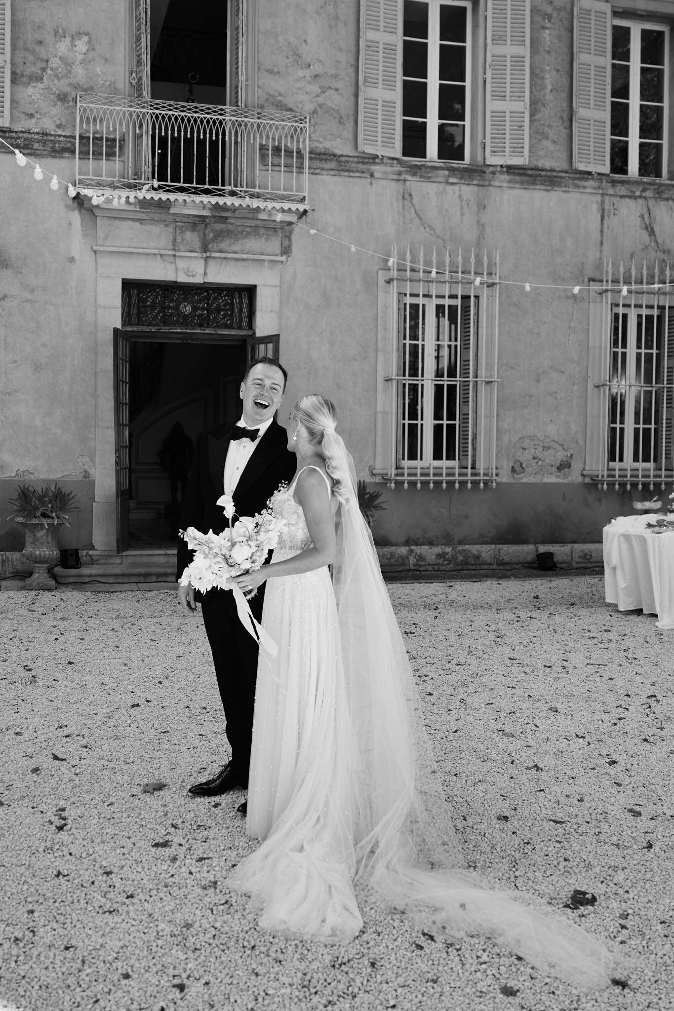 Black and white portrait of laughing couple on gravel courtyard with chateau and globe lights
