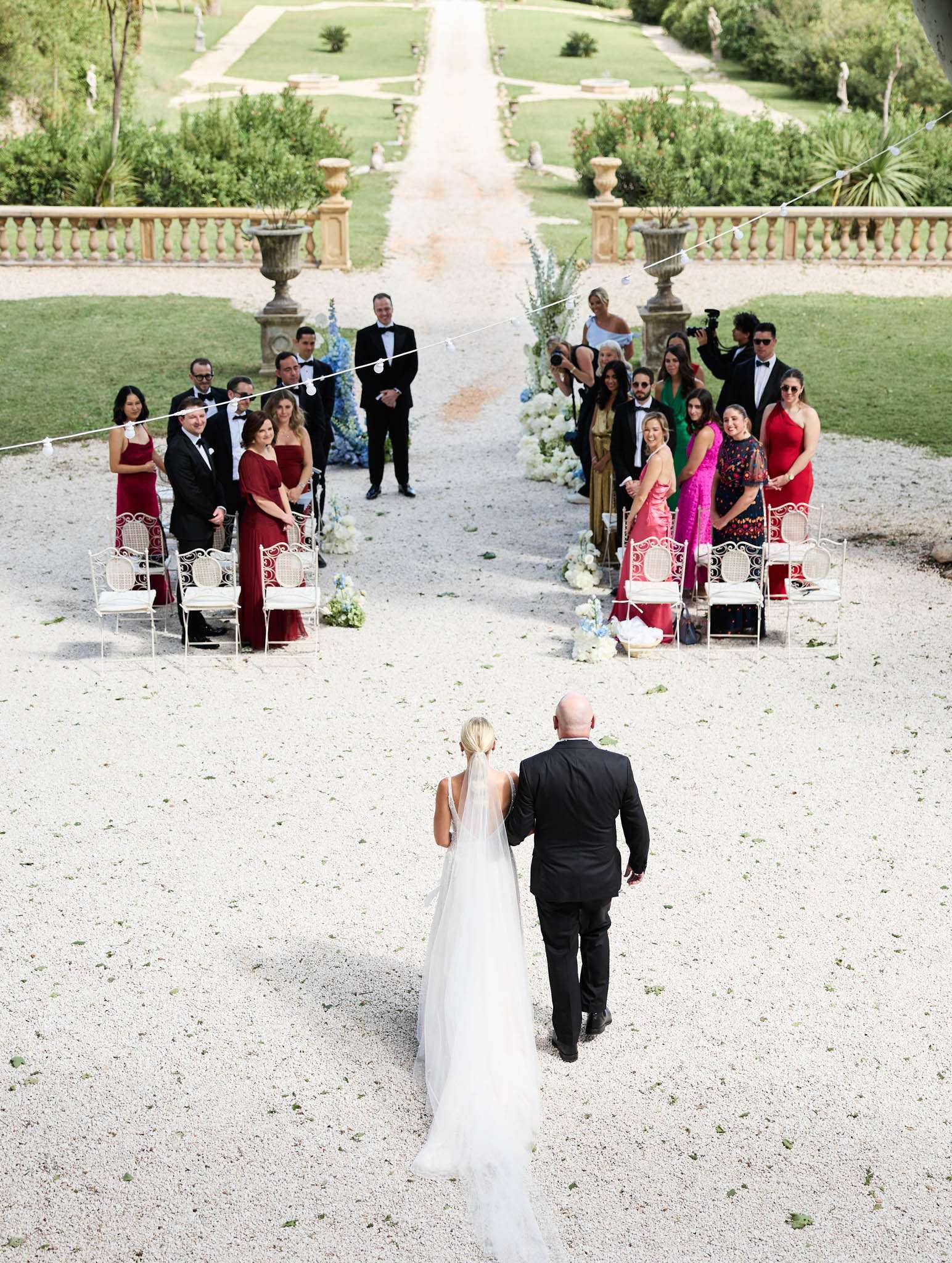 An outdoor wedding ceremony processional captured from an elevated wide-angle perspective, showing the bride walking down the aisle escorted by a companion, both viewed from behind. The bride wears a white gown with a long cathedral veil and has her blonde hair styled in a low updo; her escort wears a black suit. The ceremony takes place on a gravel path lined with white iron bistro-style chairs with cream cushions, set within the formal grounds of what appears to be a French château or villa, with a stone balustrade, large ornamental urns, and a manicured garden with a long allée stretching into the background. Approximately 20–25 standing guests flank the aisle, dressed in formal attire including burgundy and red gowns, a fuchsia dress, a gold dress, and black tuxedos. The aisle is flanked by tall floral arrangements featuring white hydrangeas, light blue delphiniums, and white blooms. The groom stands at the altar ahead, also in a black tuxedo. Light string lines are visible overhead. The decor palette combines white, soft blue, and deep red, giving a classic, formal feel.