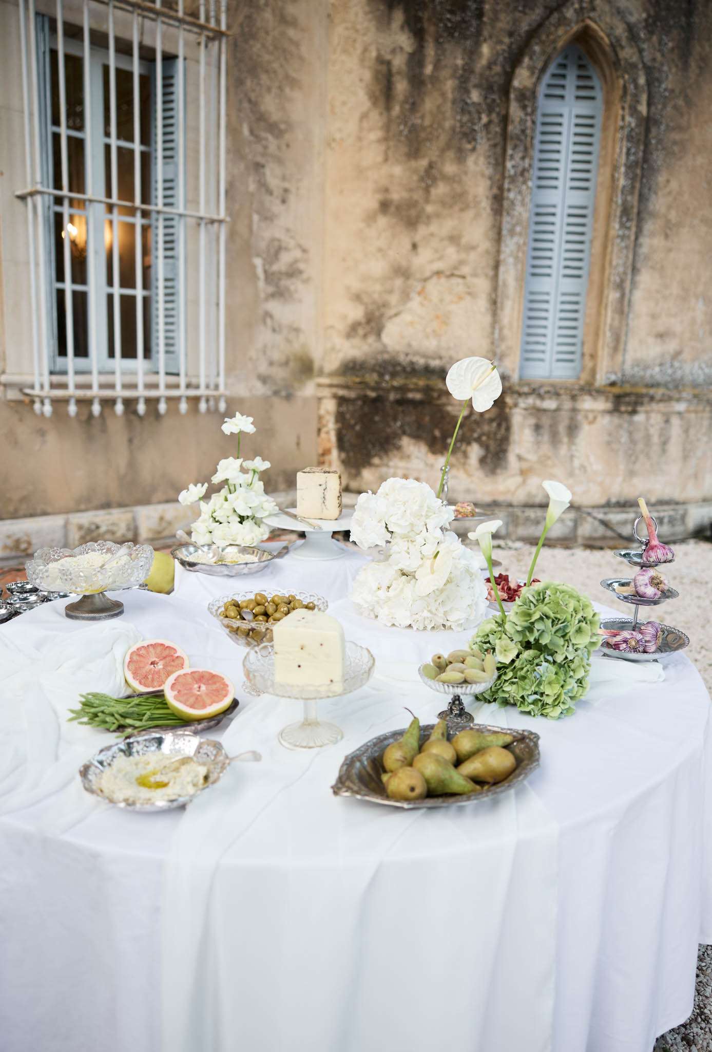 An outdoor cocktail hour or grazing table display set against the weathered stone facade of what appears to be a French château or manor house with pale blue shuttered windows. The round table is covered in a white linen and styled with an assortment of foods presented on silver platters and crystal pedestal stands, including whole cheeses, green olives, halved pink grapefruits, green beans, pears, a dip with olive oil, and garlic bulbs on a tiered silver stand. Floral arrangements in an all-white palette feature white hydrangeas, white anthurium, white calla lilies, and small white blooms, accented by clusters of green hydrangea. The overall styling is classic French with a natural, produce-forward aesthetic mixing silver serveware with crystal cake stands on a white linen cloth. Wide shot taken from a slightly elevated angle.