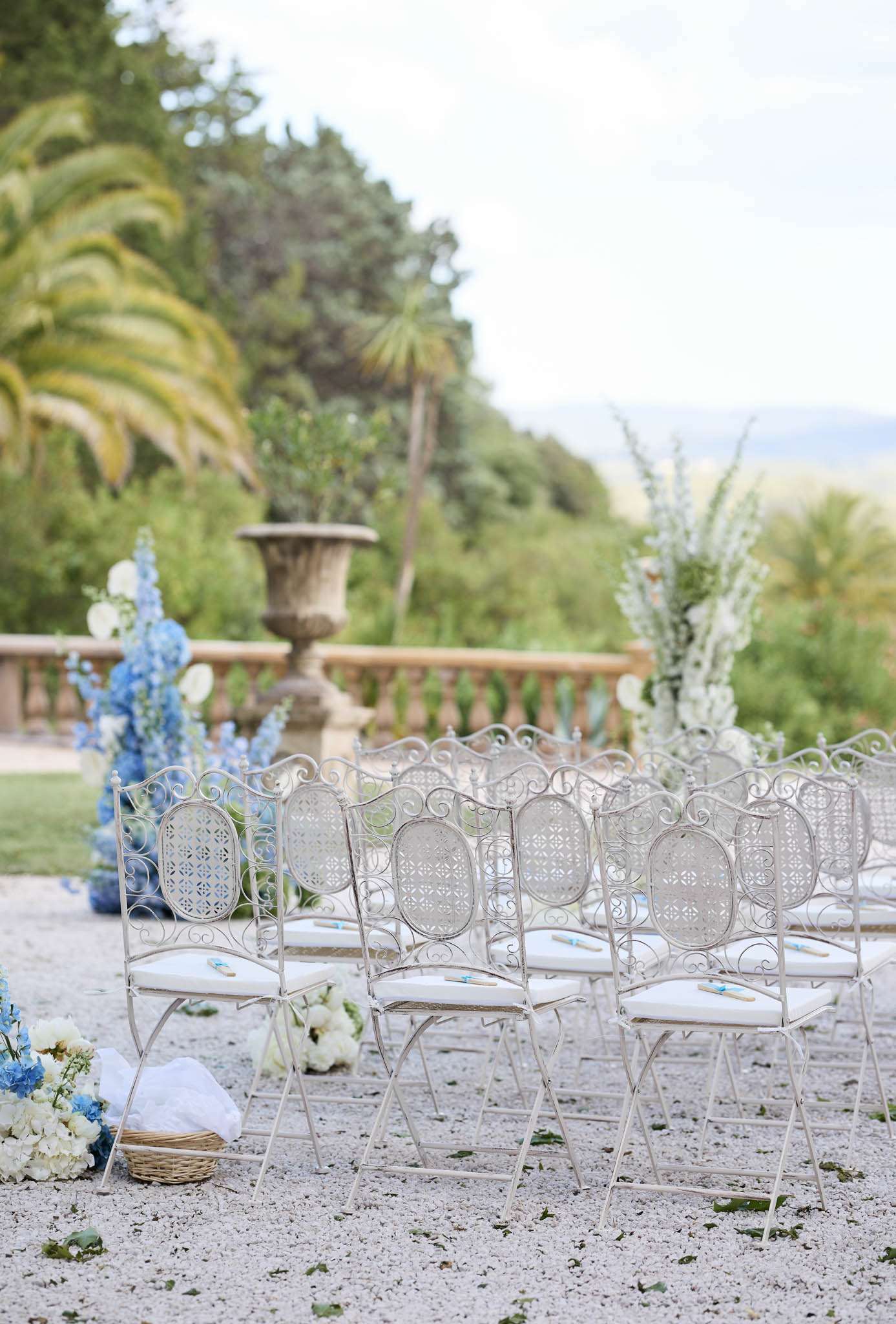 Outdoor ceremony setup with white chairs and blue delphinium aisle flowers on a chateau gravel terrace
