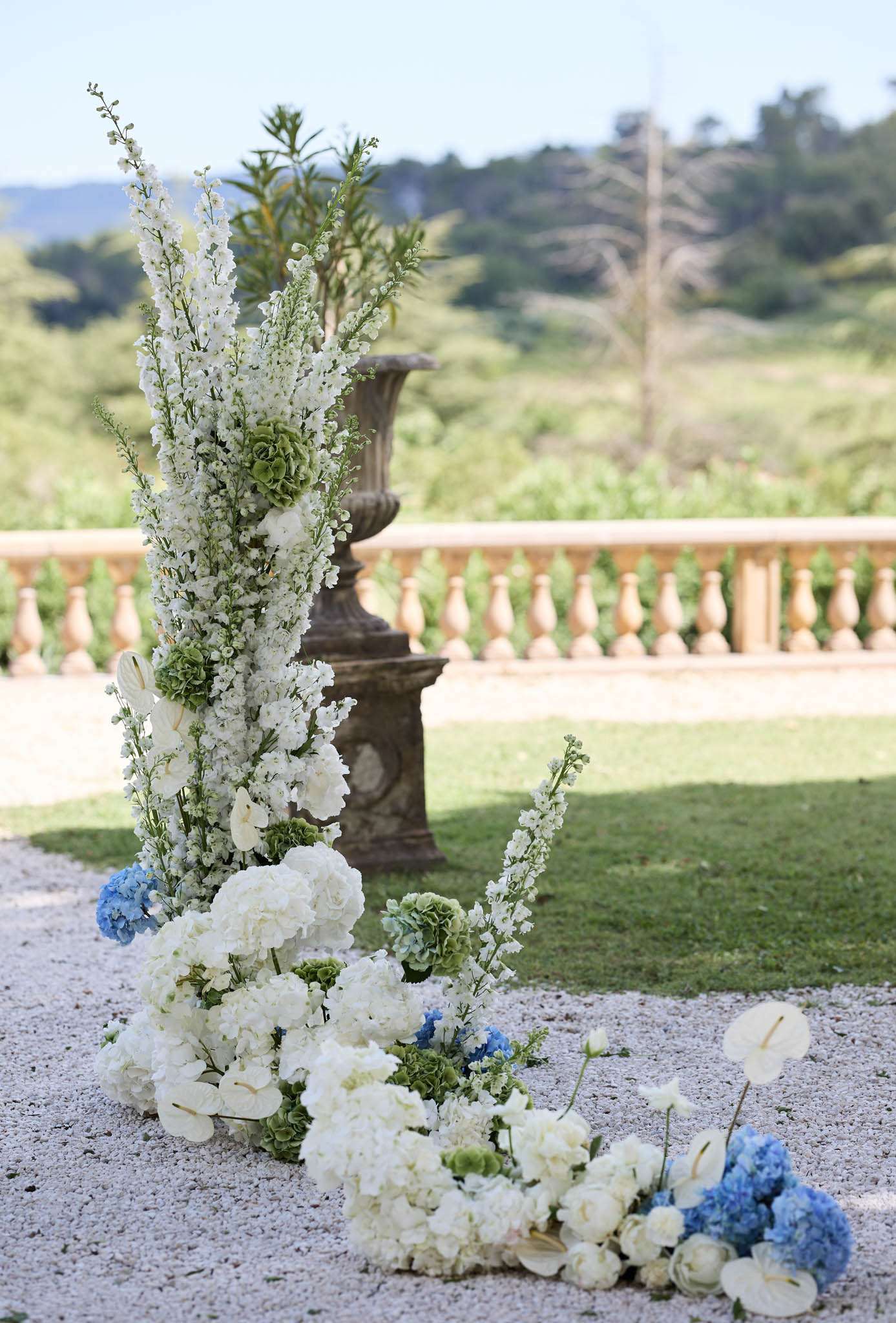 A close-up detail shot of an outdoor ceremony floral installation placed on a gravel terrace. The arrangement features tall white delphiniums and white stock as the vertical elements, accented with green hydrangeas and green viburnum, with a ground-level cluster of white hydrangeas, blue hydrangeas, white ranunculus, white anthurium, and additional blue hydrangea blooms trailing outward. The color palette is white, soft blue, and green. In the background, a stone balustrade with a large classical carved urn planter is visible, characteristic of a French château or formal garden setting. No people are present. The composition is a medium close-up shot focused on the floral structure with the venue's architectural details softly out of focus behind it.