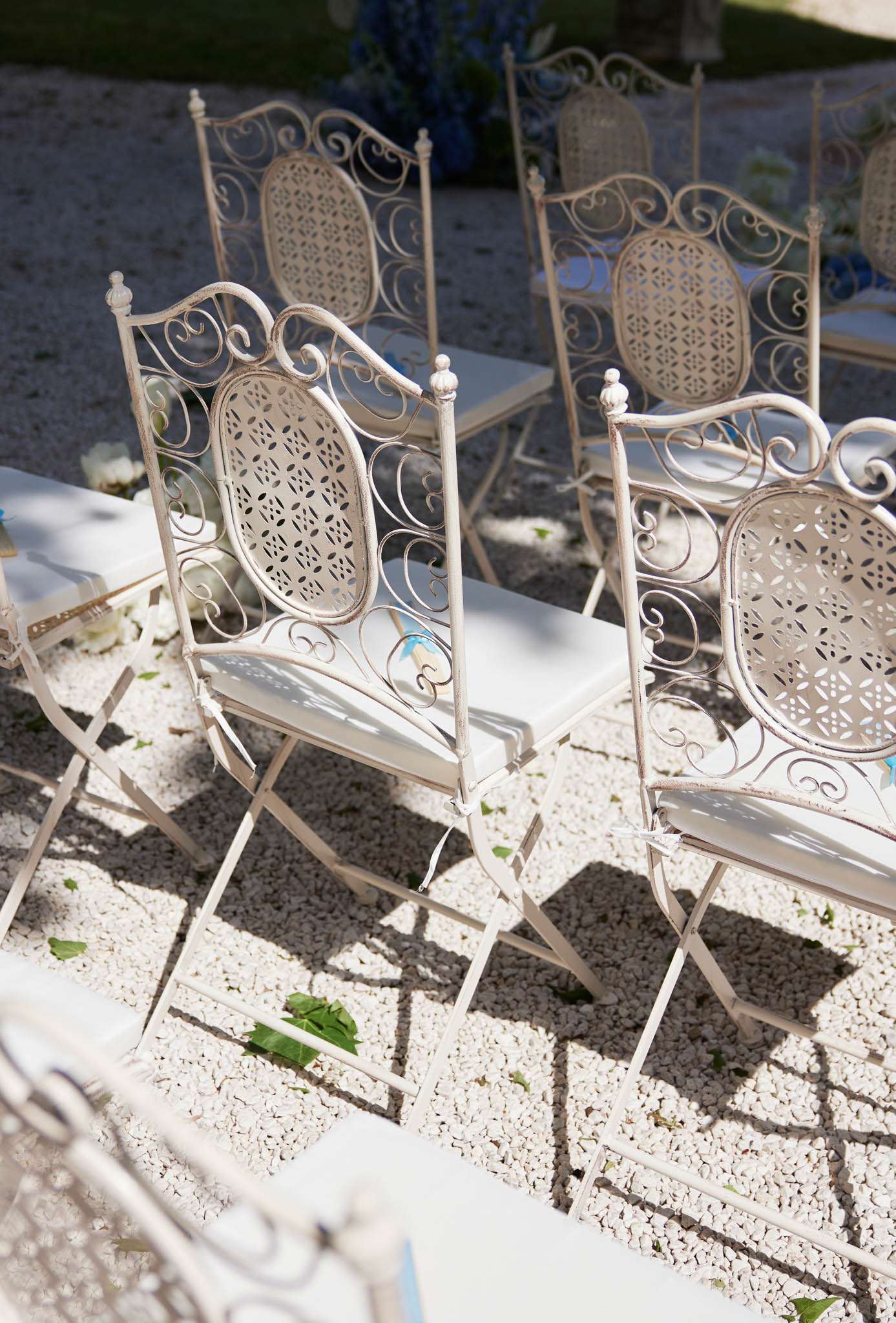 A detail shot of outdoor wedding seating arranged on a white gravel surface. The chairs are ornate cream-painted wrought iron folding chairs with intricate scrollwork and pierced oval medallion backs, fitted with white seat pads. Several chairs are visible in the frame, set around white rectangular tables. In the background, partially visible floral arrangements in blue tones — likely hydrangeas or delphiniums — are displayed in cobalt blue vessels. The setting appears to be an outdoor ceremony or cocktail area with a classic French provincial aesthetic. The image is taken from a slightly elevated angle in bright natural light.