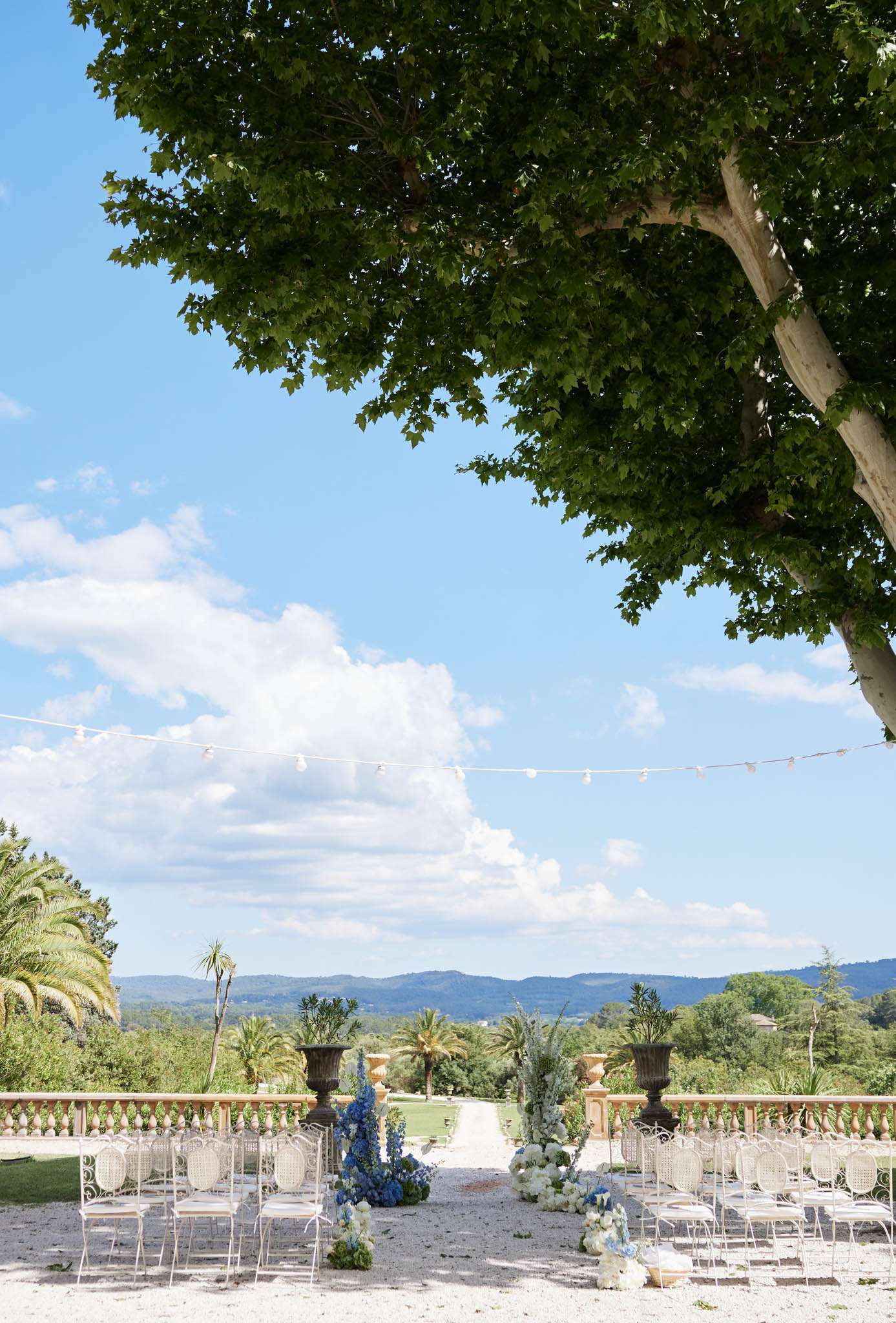 An outdoor wedding ceremony setup on a gravel terrace with an open panoramic view of rolling hills and gardens in the background. The aisle is flanked by white metal chiavari-style chairs arranged in rows, with floral arrangements featuring tall blue delphiniums, white blooms, and greenery placed at the aisle ends alongside dark cast-iron urns on pedestals. String lights with globe bulbs are strung overhead across the space, and a stone balustrade runs along the terrace edge. The styling palette is white and blue, with a classic French estate aesthetic. Wide shot capturing the full ceremony layout and surrounding landscape. Potential venue feature image.