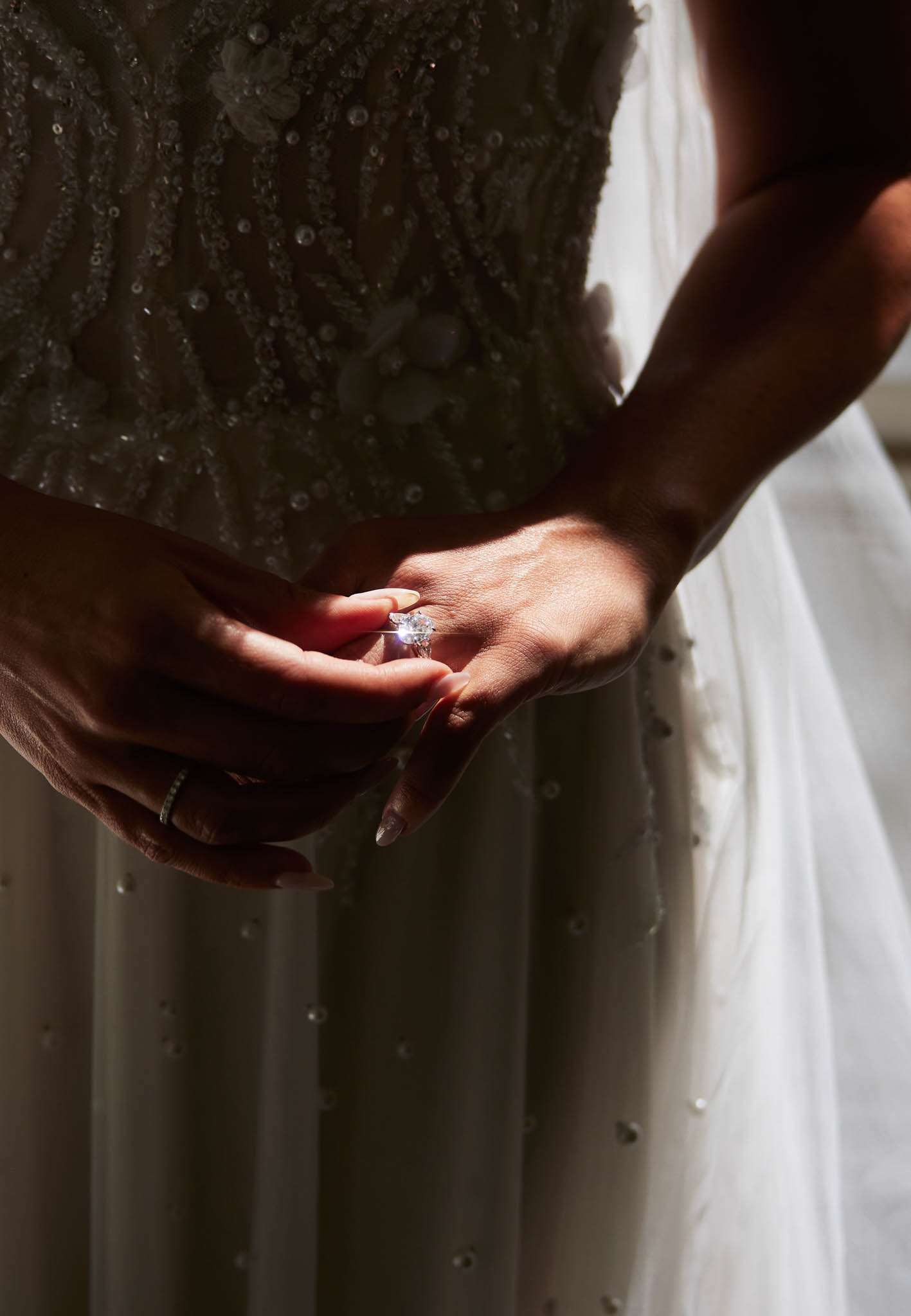 Close-up of oval diamond engagement ring catching light on bride's hand over beaded gown