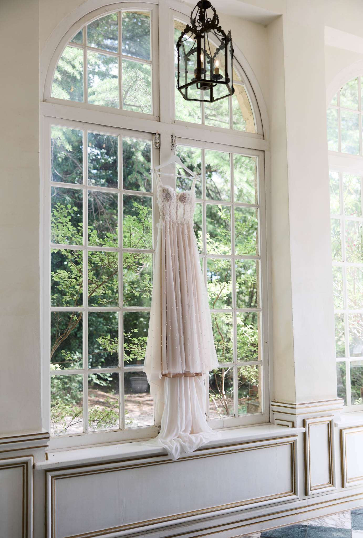 Blush-ivory bridal gown with lace bodice and pearl embellishments hanging before arched window with gold trim