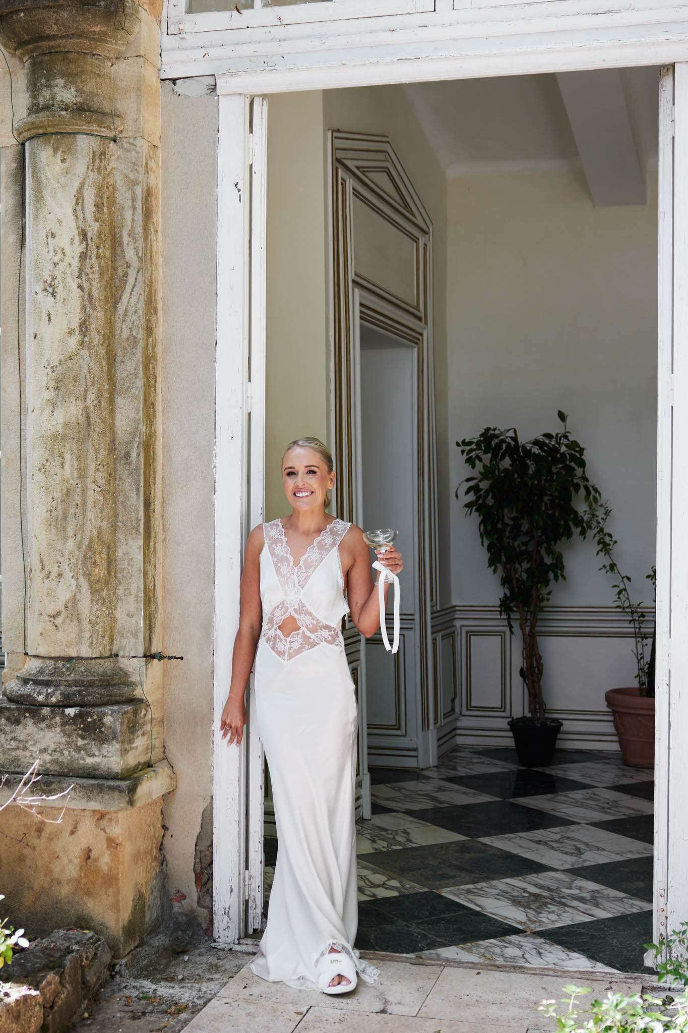 A bride stands relaxed in the open doorway of a French château, leaning against the door frame and holding a champagne coupe with a white ribbon tied around the stem. She wears a sleek ivory slip-style wedding gown with deep lace V-panel inserts at the bodice and a fitted silhouette, paired with white open-toe sneakers. Her hair is pulled back in a low, sleek updo. Behind her, the interior features black-and-green marble chequerboard flooring, ornate white-and-gold panelled walls, and a large potted ficus tree. The exterior shows a weathered stone column with peeling white-painted architectural detailing, suggesting a classic French country estate. Full-length portrait shot taken from a slight low angle.