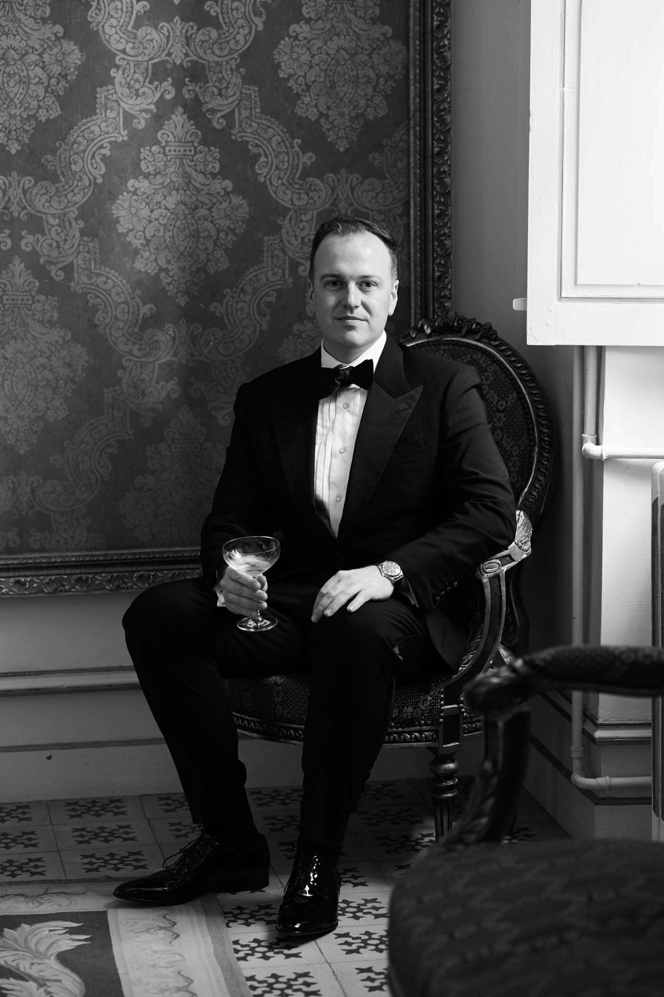 Black-and-white portrait of a groom seated in an ornate carved armchair inside what appears to be a château or manor house interior. He is dressed in a black tuxedo with a white dress shirt featuring button studs and a black bow tie, paired with polished black Oxford shoes and a silver dress watch. He holds a coupe or martini-style cocktail glass in one hand. The background features heavily patterned damask wallpaper and a large gilt-framed artwork, while the floor displays decorative patterned cement tiles. The image uses strong contrast with deep blacks and bright highlights, giving it a classic, editorial feel. Full-length seated portrait composition shot indoors.