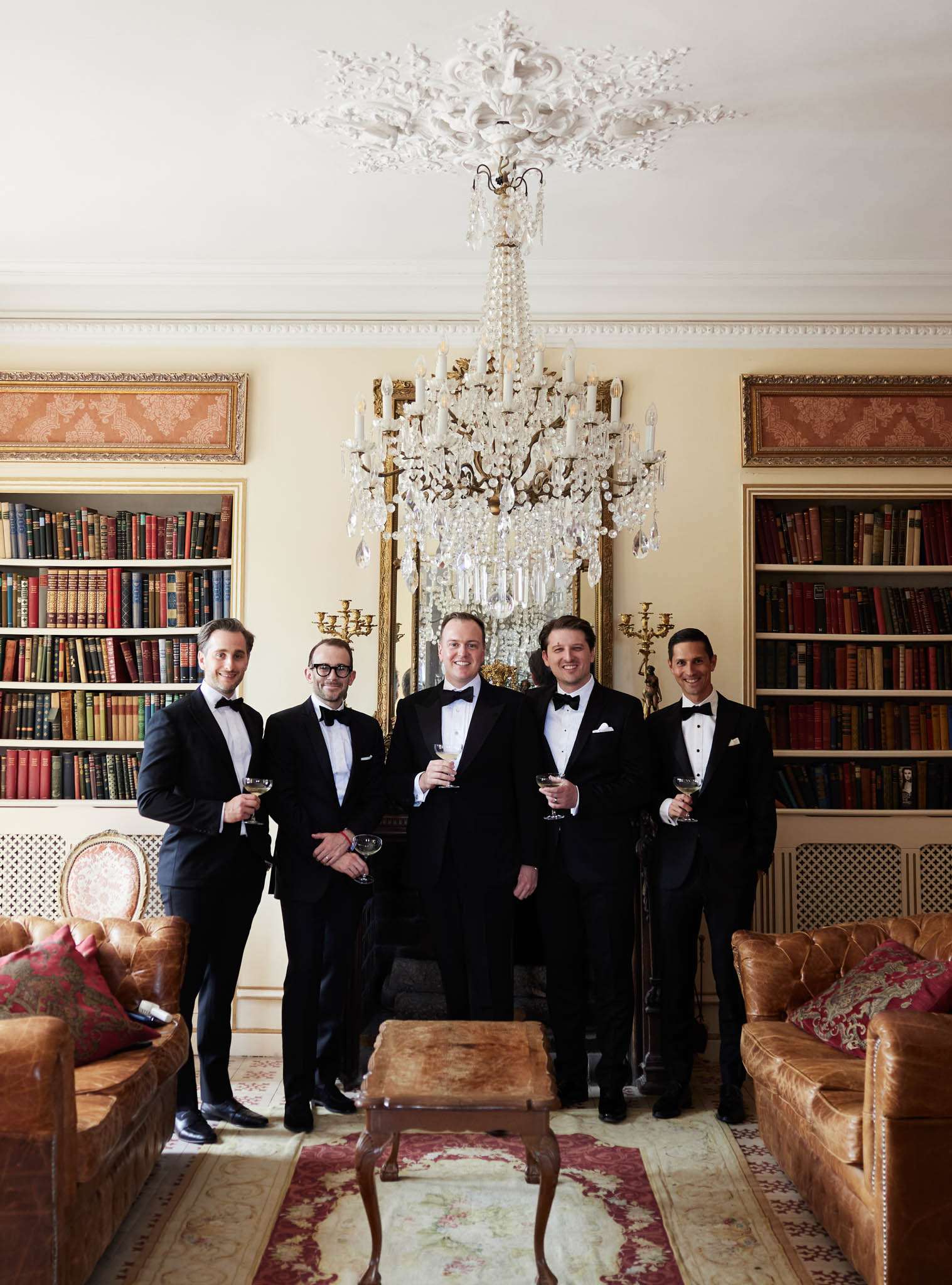 Five men in tuxedos with champagne coupes in chateau library with leather-bound books and crystal chandelier