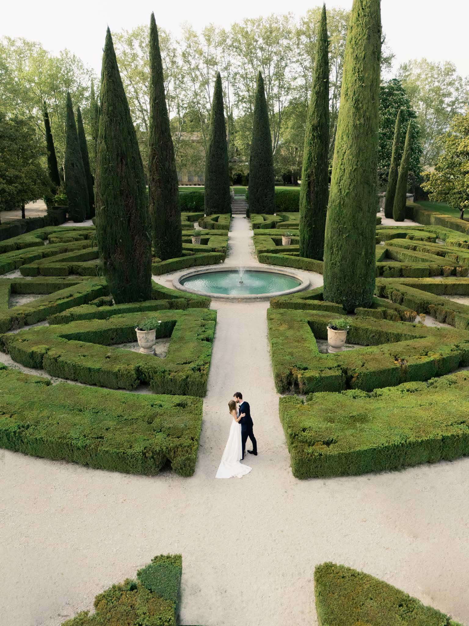 Aerial view of bride and groom in formal French parterre garden with boxwood hedges and stone fountain