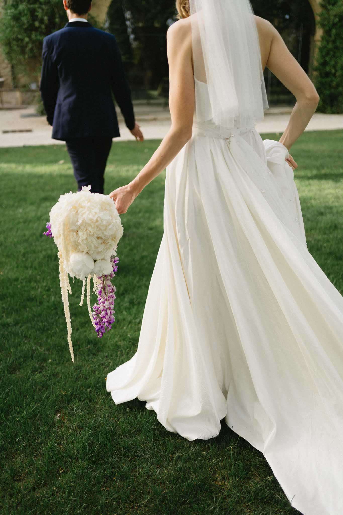 Bride and groom walking away across lawn, bride holding peony bouquet with trailing lupin stems