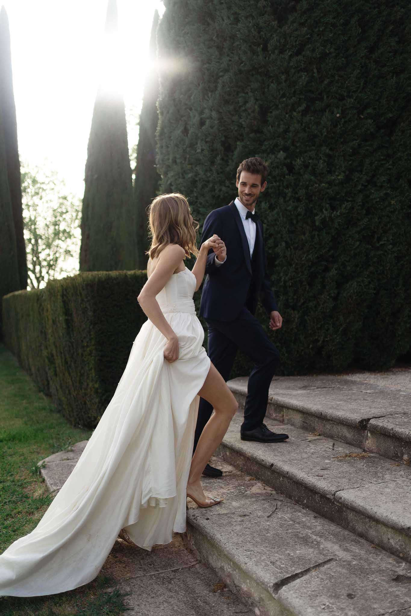 Bride and groom walking up stone steps between cypress topiaries in golden hour backlight