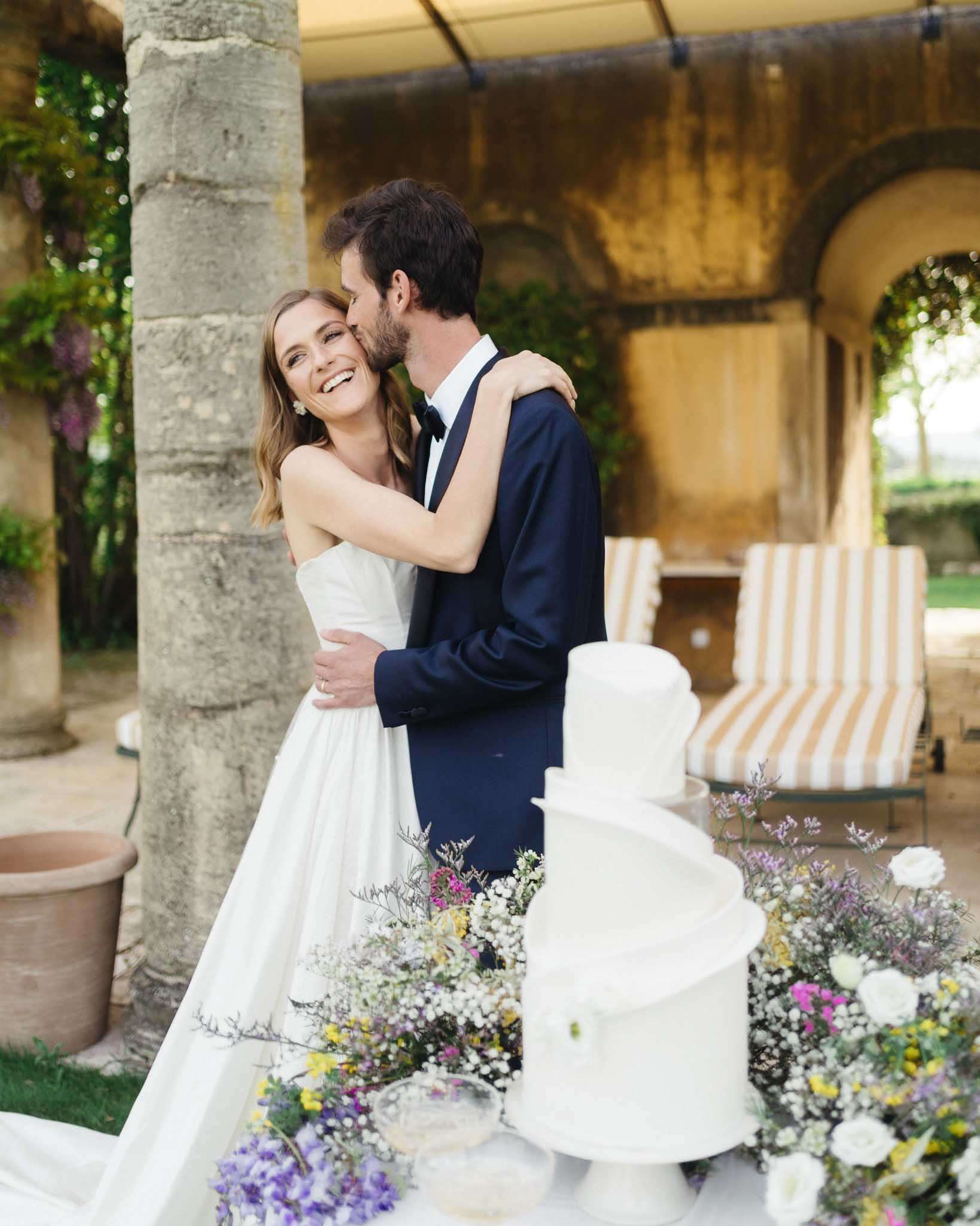 A couple portrait taken outdoors on a covered terrace or loggia with stone arched columns, likely at a French or Italian château or villa. The bride wears a strapless white A-line gown with a full skirt and short wavy hair with pearl earrings, smiling broadly as the groom kisses her cheek. The groom is dressed in a navy blue suit with a black bow tie. In the foreground, a three-tiered smooth white fondant wedding cake is displayed on a table surrounded by loosely arranged wildflower clusters in purple, yellow, hot pink, white gypsophila, and lavender sprigs. Striped cream and tan lounge chairs are visible in the soft background. The shot is a medium couple portrait with the cake styled as a foreground detail element.