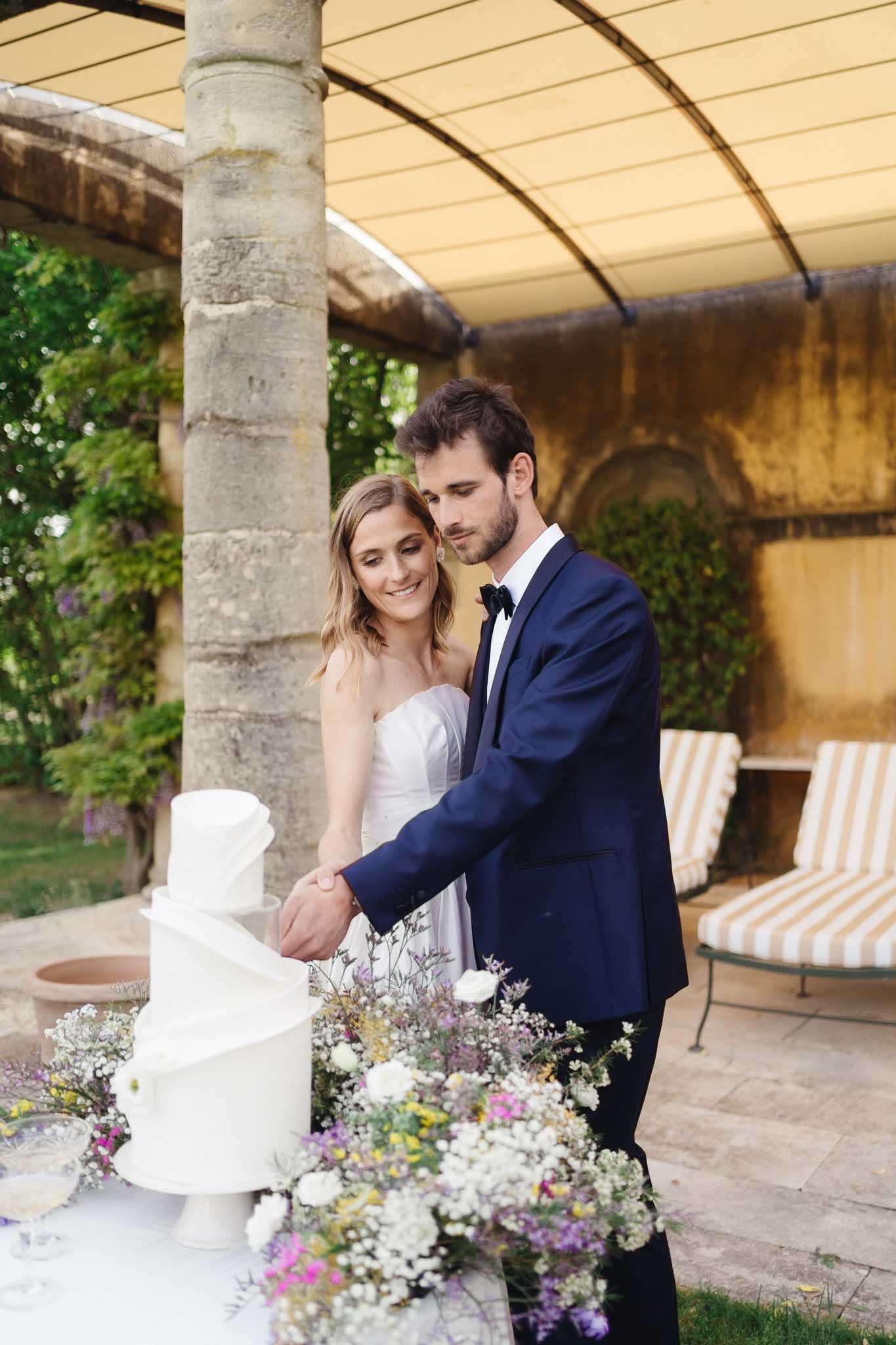 Bride and groom cutting three-tier white wedding cake decorated with wildflowers under stone colonnade on outdoor terrace