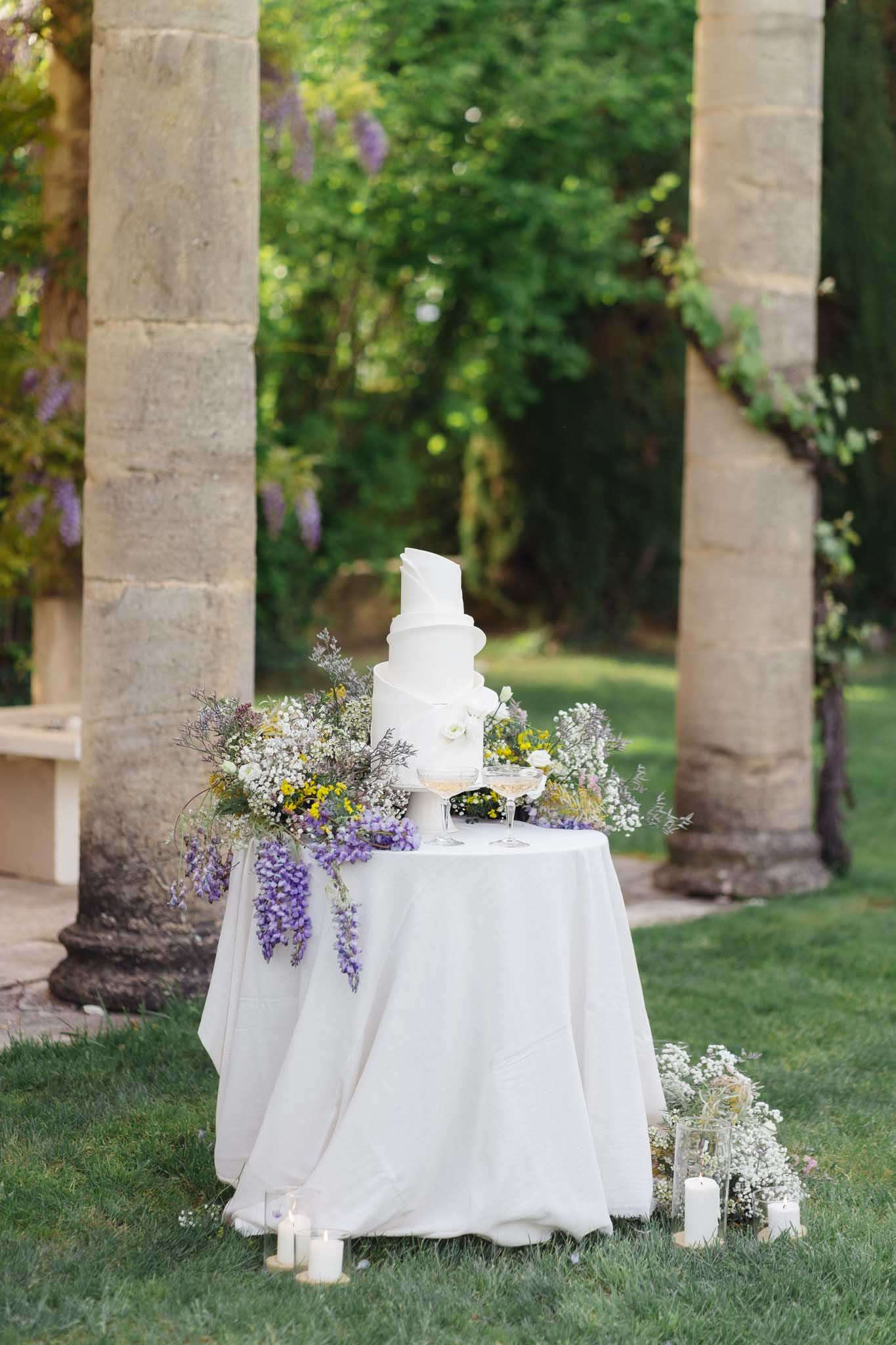Four-tier white draped cake with wisteria, baby's breath, and yellow wildflowers between stone columns