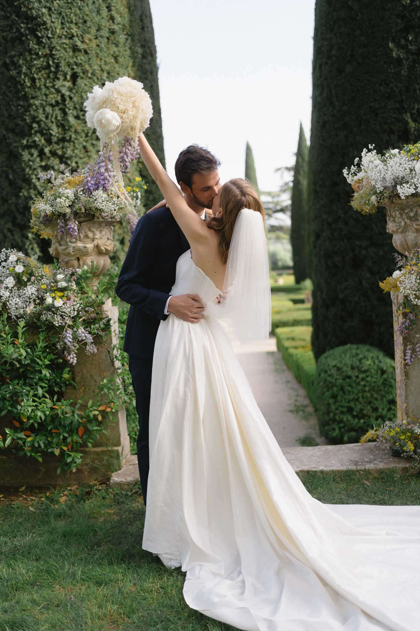 Couple kissing with bouquet raised between cypress trees and wisteria-draped stone urns in formal garden