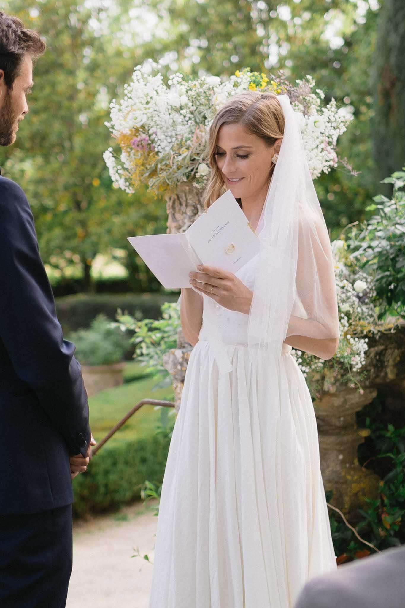 Bride reading personal vows from a booklet during an outdoor garden ceremony with floral urn behind