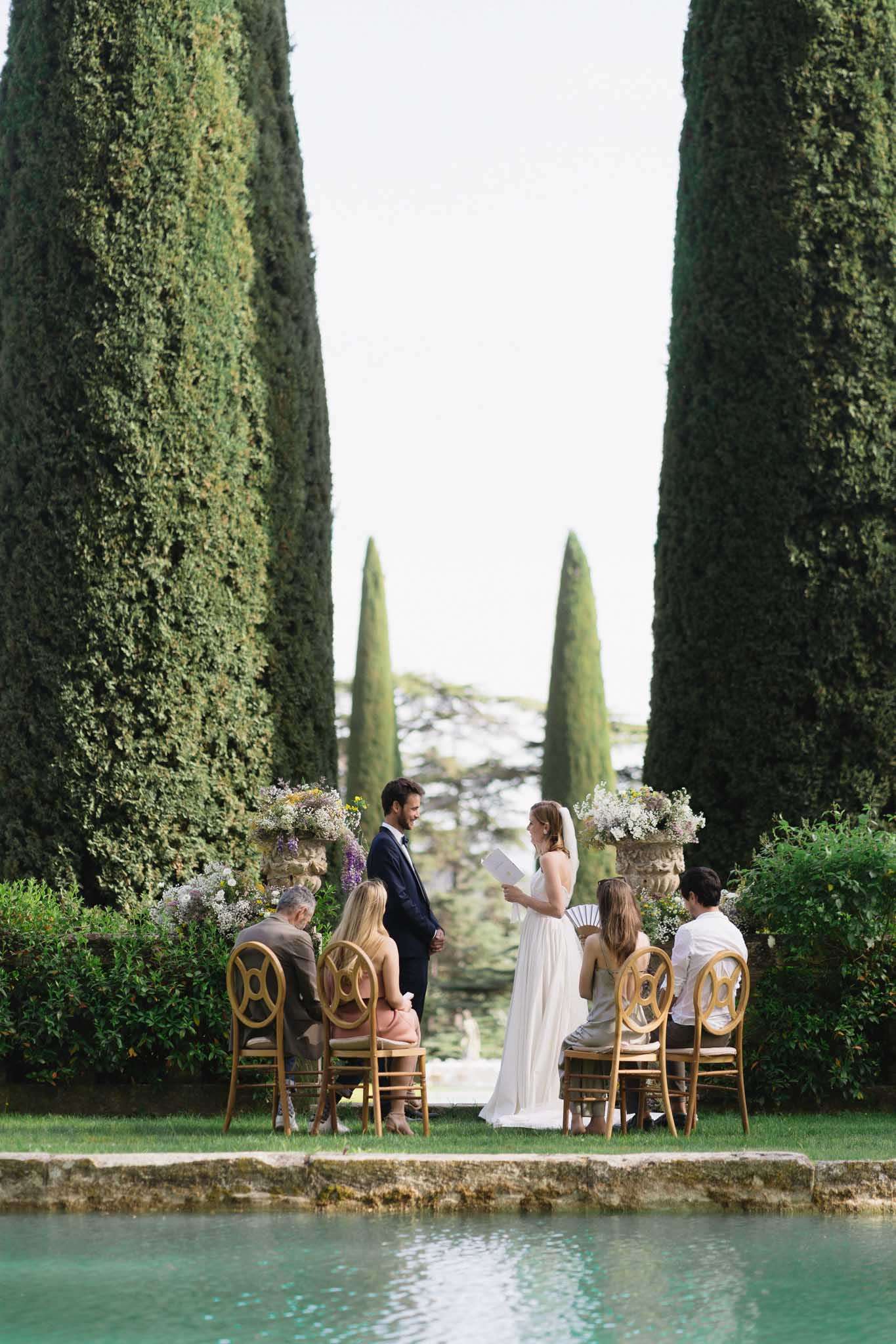 Couple exchanging vows in formal garden with stone urns of wildflowers, viewed across ornamental pool with cypress trees