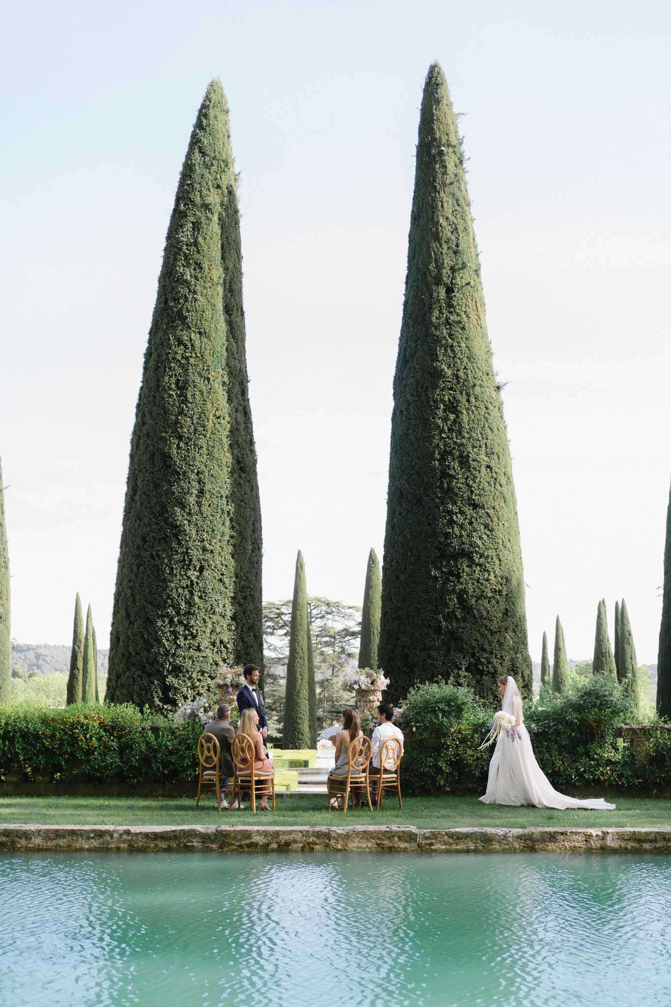 An outdoor wedding ceremony taking place in a formal garden setting, photographed from across a rectangular ornamental pool with turquoise-tinted water. The ceremony is intimate, with approximately five guests seated on gold Chiavari-style chairs arranged in a small semicircle, facing a groom in a dark suit. The bride stands to the right in a white gown with a long cathedral-length veil and carries a bouquet featuring what appears to be white and purple flowers, including lavender or wisteria. Floral arrangements in soft, muted tones — likely cream, blush, and lilac — are displayed in large decorative urns flanking the ceremony space. The composition is a wide shot that emphasizes the dramatic landscape framing, with two very tall, narrow cypress trees rising symmetrically behind the ceremony and a row of additional cypress trees extending into the background. Potential venue feature image.