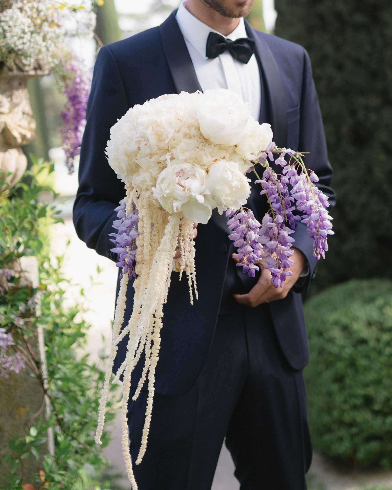 A groom in a navy tuxedo with satin lapels and a black bow tie holds a large cascading bridal bouquet in an outdoor garden setting. The bouquet features cream peonies at the top, trailing ivory amaranthus, and clusters of purple wisteria draping downward in a dramatic waterfall shape. The composition is a mid-shot portrait focused on the torso, with the groom's face partially cropped at the top of the frame. The floral design has a classic yet abundant style, with the purple and cream color palette contrasting sharply against the dark navy of the tuxedo.
