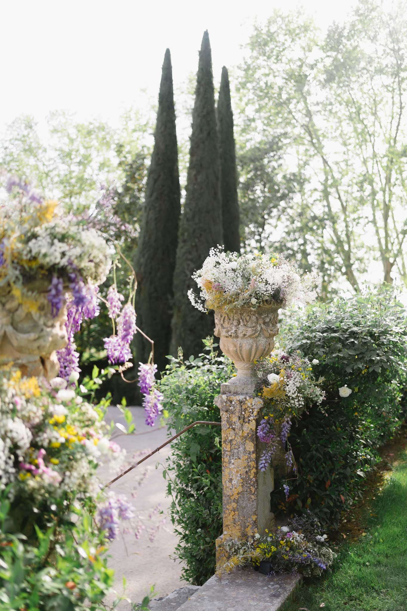 Stone urn with gypsophila, cream roses, and purple wisteria alongside floral garden arch