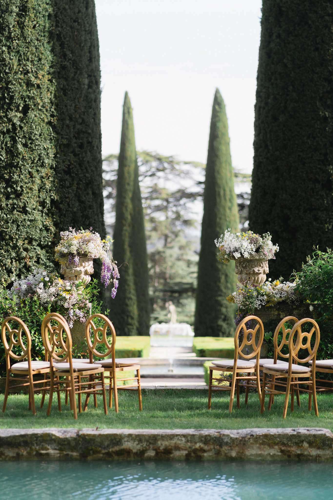 Ceremony with gold chairs and lavender-cascading stone urns before cypress-lined garden alley