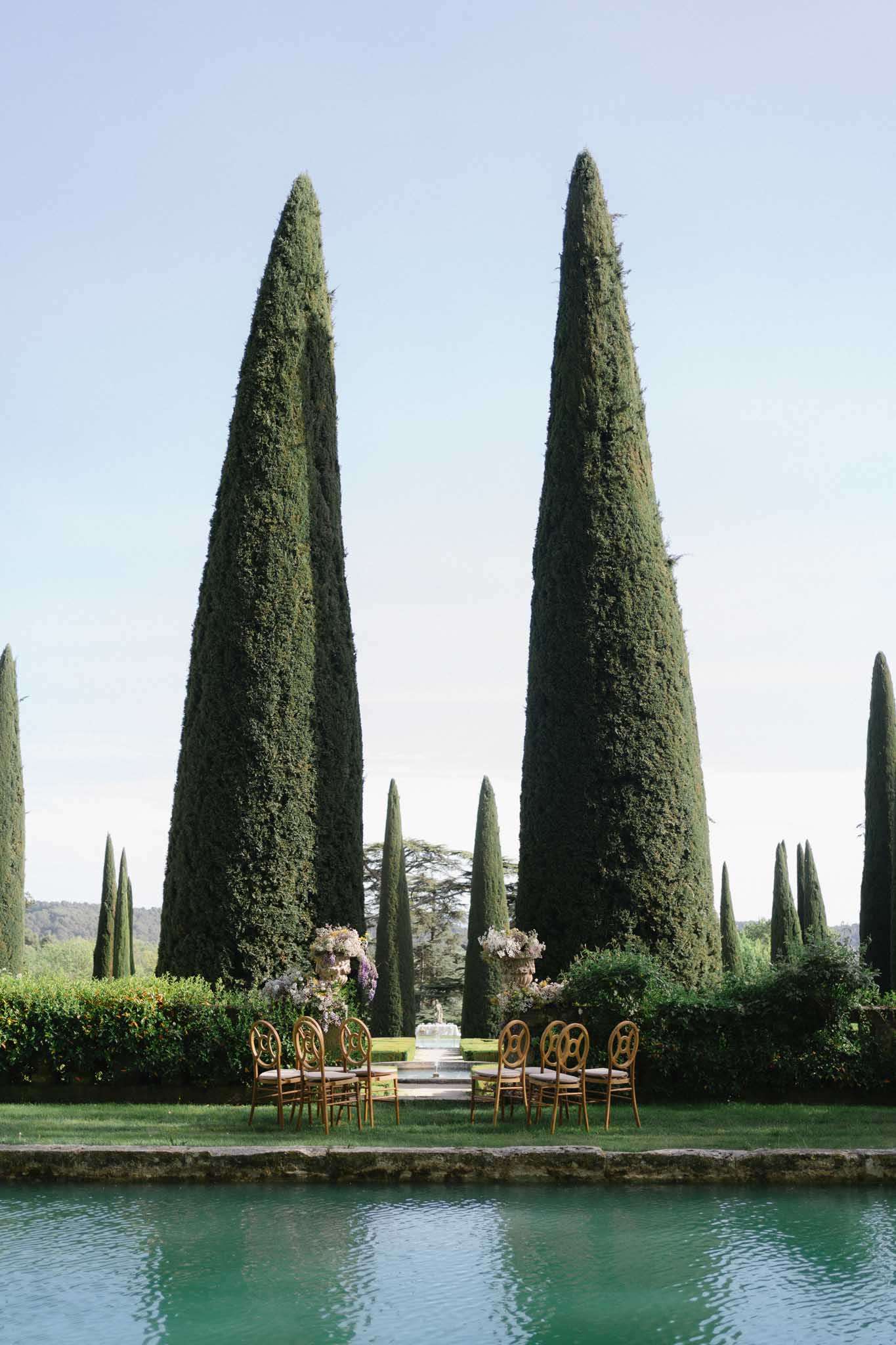 Intimate ceremony setup with gold chairs and pink floral urns beside ornamental pool framed by cypress trees