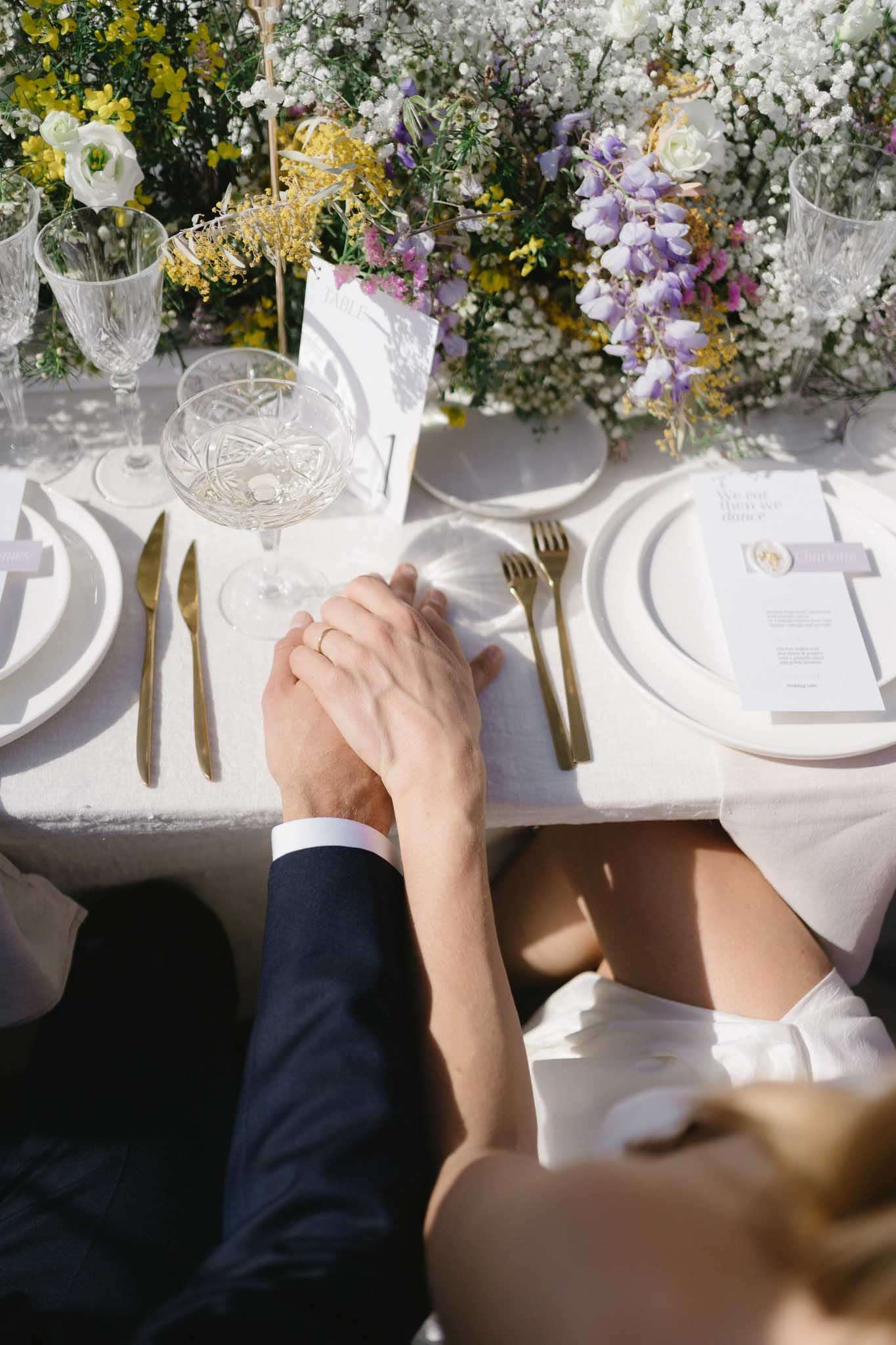 Overhead couple holding hands at table with wildflower centerpiece of wisteria mimosa and baby breath