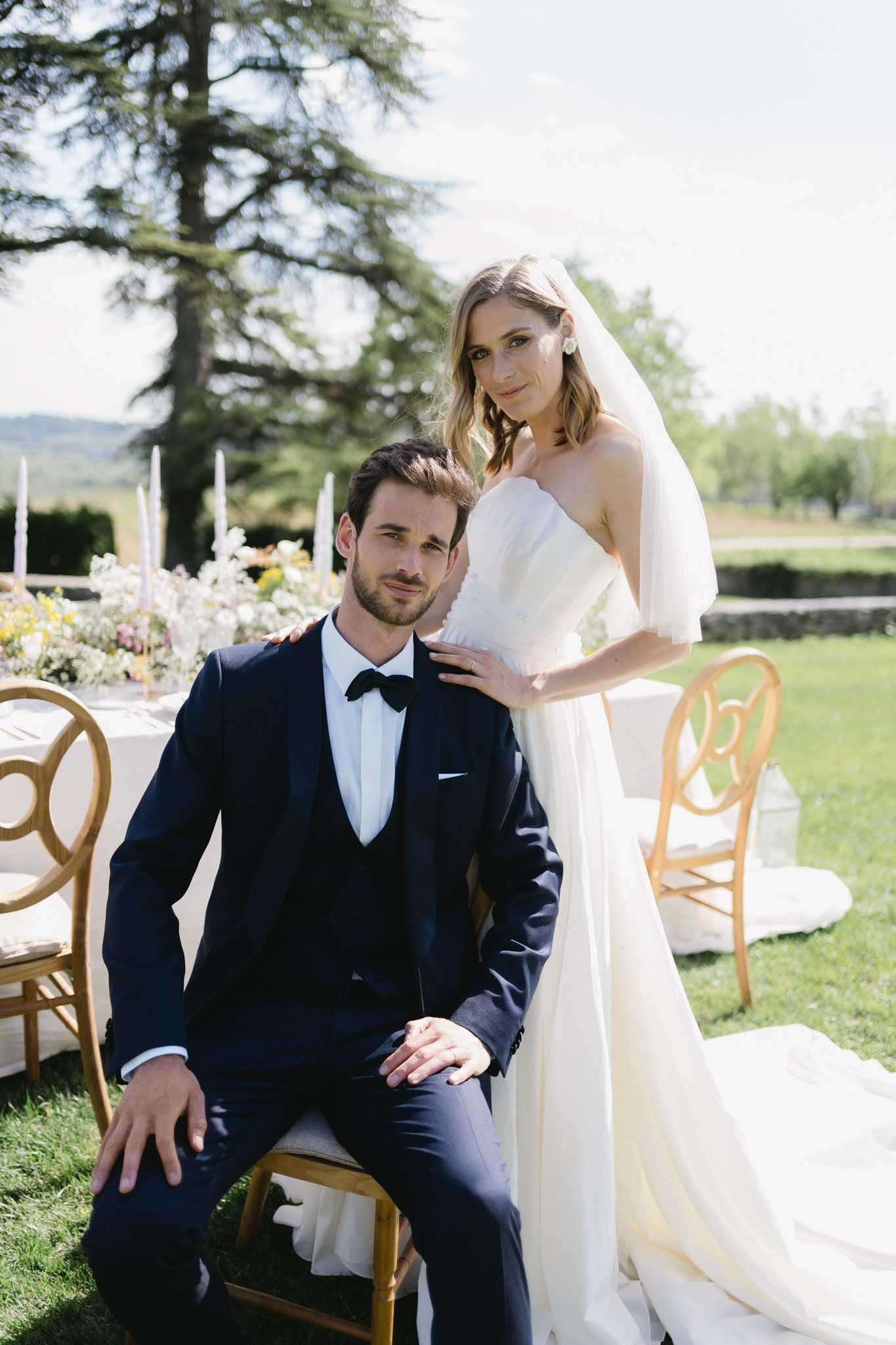 A couple portrait taken outdoors at what appears to be a garden reception setup, with the groom seated on a gold Chiavari chair and the bride standing behind him with her hand resting on his shoulder. The groom wears a navy three-piece suit with a black bow tie and white dress shirt, while the bride wears a strapless ivory gown with a flowing chiffon overlay and a long veil, her blonde hair worn loosely. In the background, a styled reception table is visible dressed in white linens with tall taper candles in blush tones and a low floral arrangement featuring white, yellow, and soft mixed blooms; gold Chiavari chairs surround the table. The setting is an open outdoor lawn area with a wide landscape view behind. The shot is a medium portrait with a slightly shallow depth of field, keeping the couple sharp against the softly blurred tablescape.