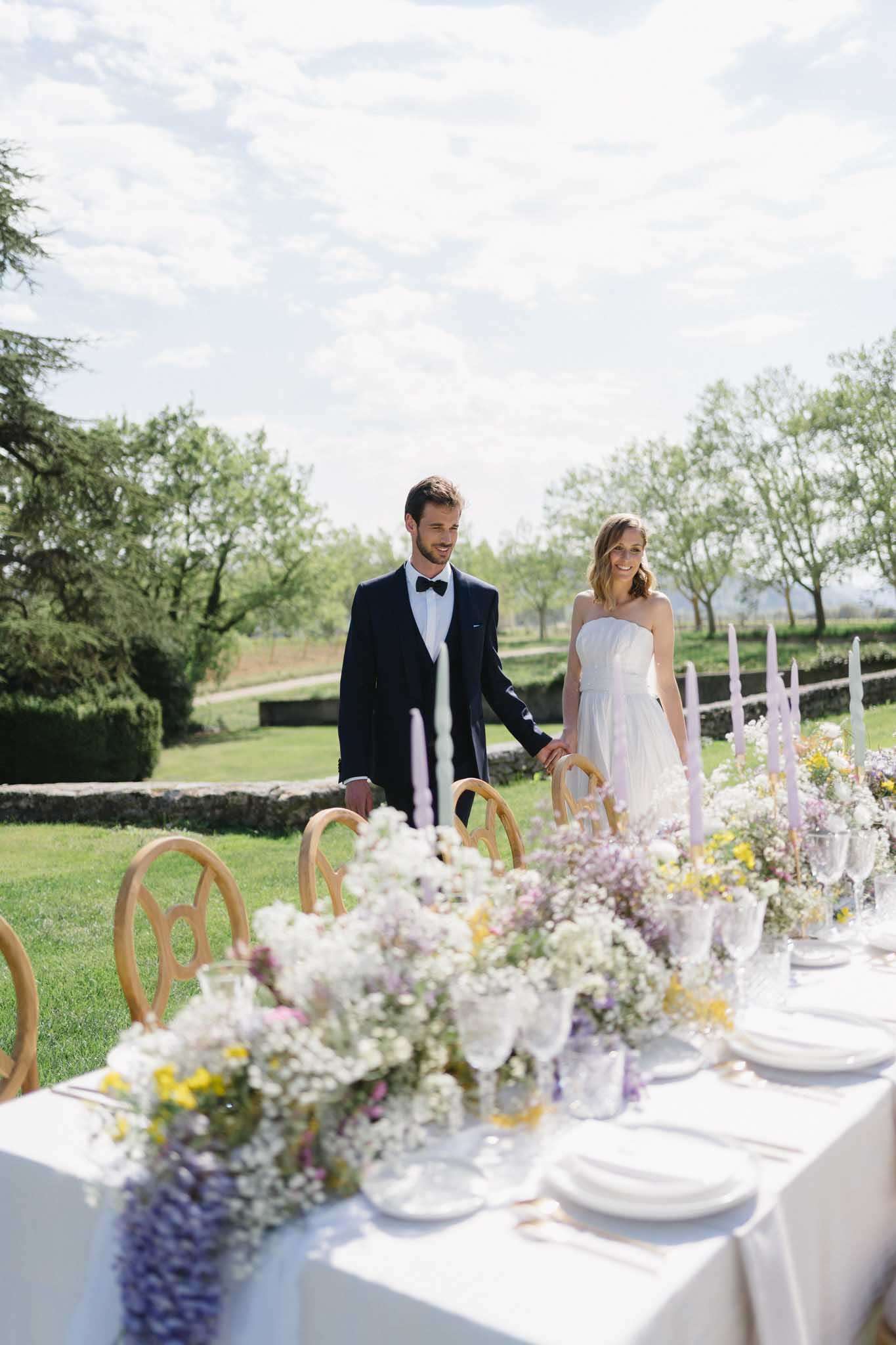 A couple stands hand-in-hand beside a styled outdoor reception table set in a garden with a low stone wall and open parkland in the background. The groom wears a navy tuxedo jacket with a black bow tie and white shirt, while the bride wears a strapless white dress with a layered skirt. The long table is covered in a white linen and features a lush floral runner composed of white gypsophila, lavender wisteria, yellow wildflowers, and soft pink blooms, giving a loose, garden-picked aesthetic. Tall tapered candles in pale lavender and blush are interspersed along the table alongside clear glassware, white plates, and small bud vases. The chairs are natural wood with a rounded back detail. The overall styling palette is soft pastels — lavender, yellow, and white — with a light, airy, garden-party feel. The shot is taken from a low angle at table level, placing the couple in the mid-ground against the outdoor setting.