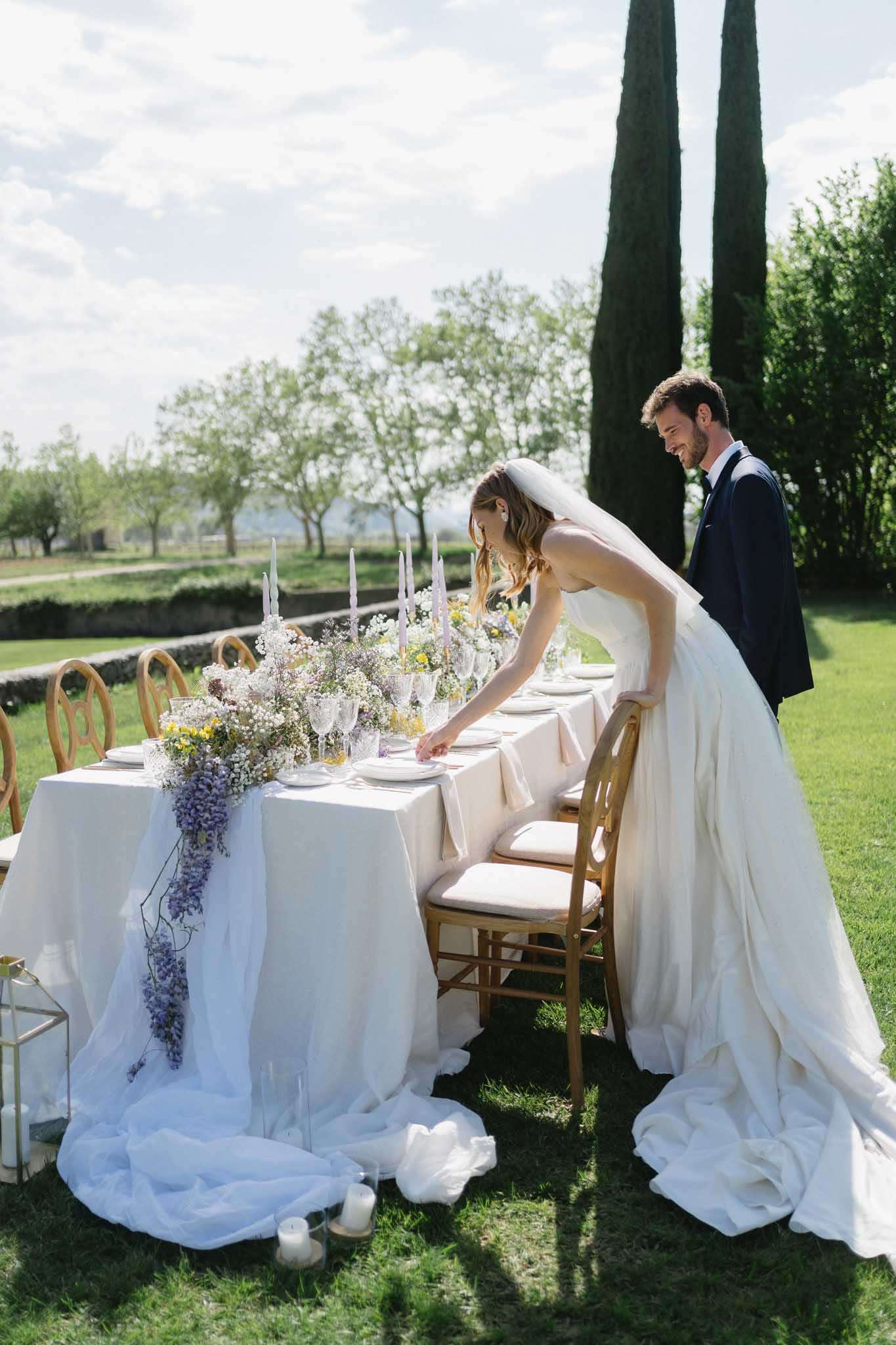 Bride and groom at outdoor reception table with lavender wisteria garland centerpiece and gold cross-back chairs