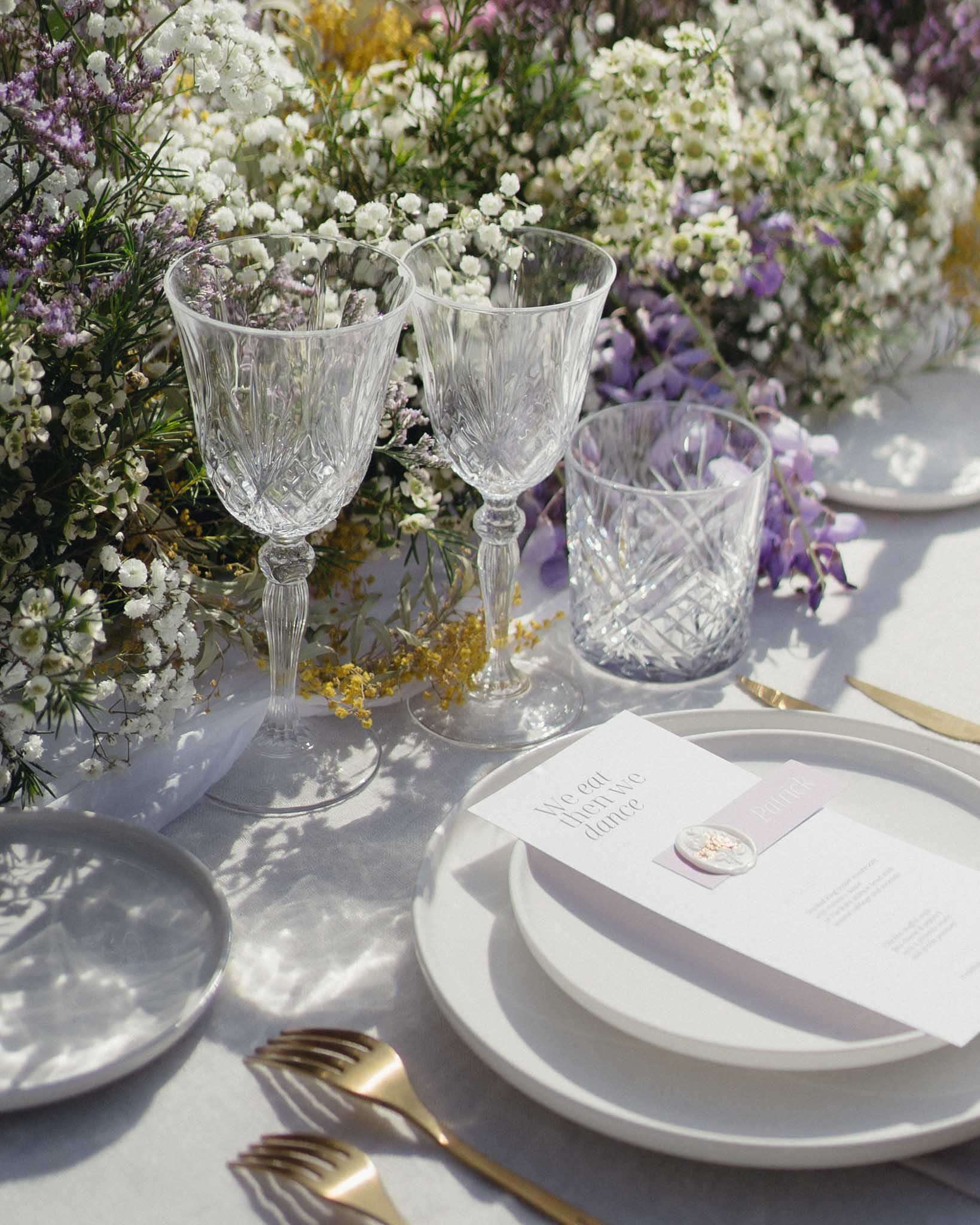 A close-up detail shot of a wedding reception table setting, photographed in bright natural light. The place setting features stacked white plates on a white linen tablecloth, accompanied by gold flatware, two cut-crystal wine glasses, and a cut-crystal tumbler. A menu card printed with the phrase 'We eat then we dance' is placed on the plates, secured with a pale lilac wax seal. Loose floral arrangements in white gypsophila, white waxflower, purple delphinium, and small yellow mimosa blooms spill across the table in an unstructured, garden-style arrangement. The overall decor palette is white, soft lavender, and gold, with a clean modern aesthetic.