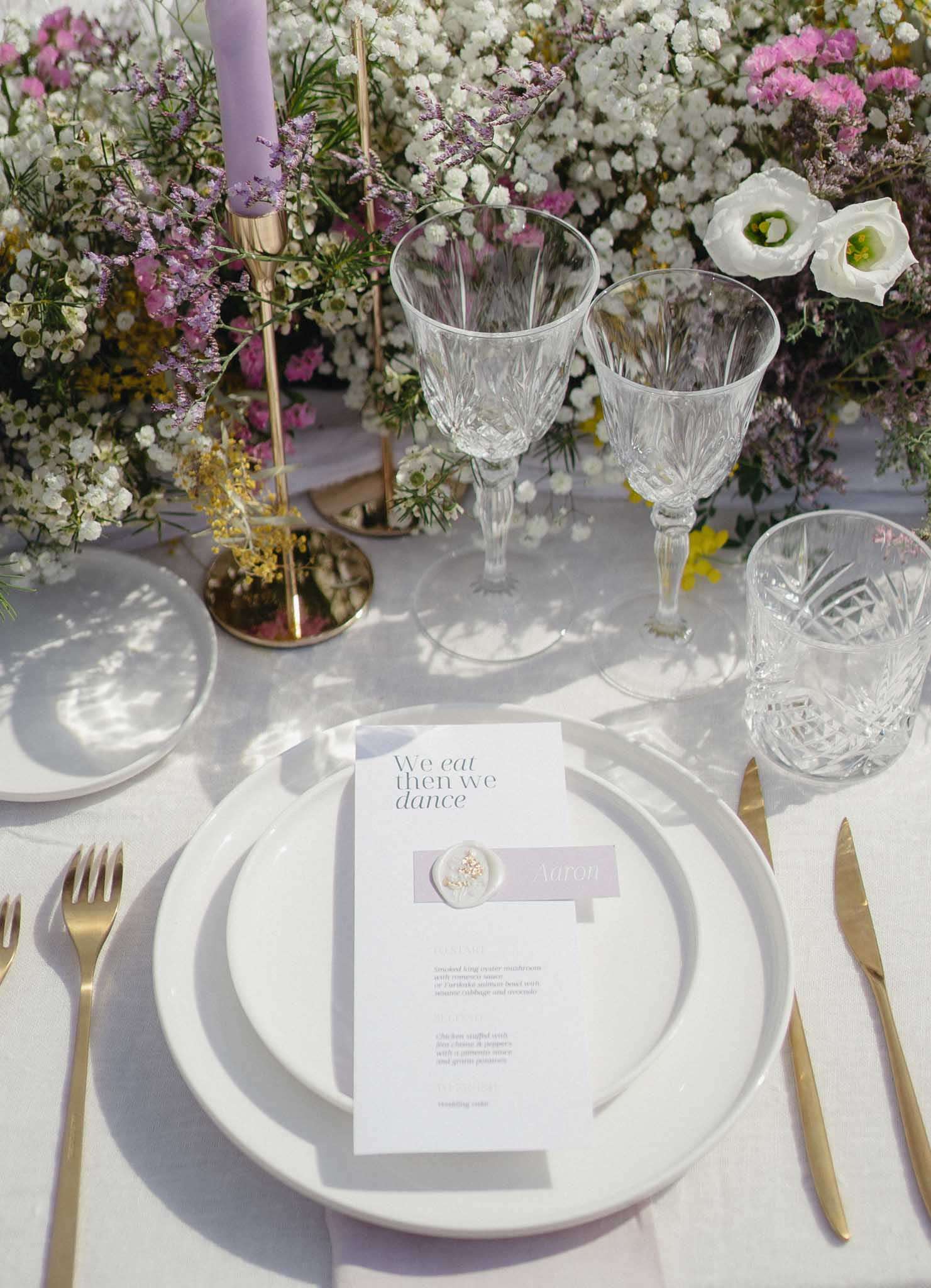 A close-up detail shot of a single wedding reception place setting on a white linen tablecloth. The place setting features stacked white ceramic plates with gold flatware (two forks on the left, knife and additional piece on the right), two cut-crystal wine glasses and one cut-crystal tumbler, and a menu card placed on the top plate. The menu card reads 'We eat then we dance' in a modern serif font with a lilac accent block bearing the guest name 'Aaron' and a small wax seal with dried florals. A tall gold taper candleholder holds a lavender-purple taper candle. The background is filled with an abundant floral arrangement featuring white gypsophila, hot pink statice, white lisianthus, yellow mimosa, and purple limonium. The overall styling palette is white, gold, lavender, and pink with a modern, garden-inspired aesthetic.