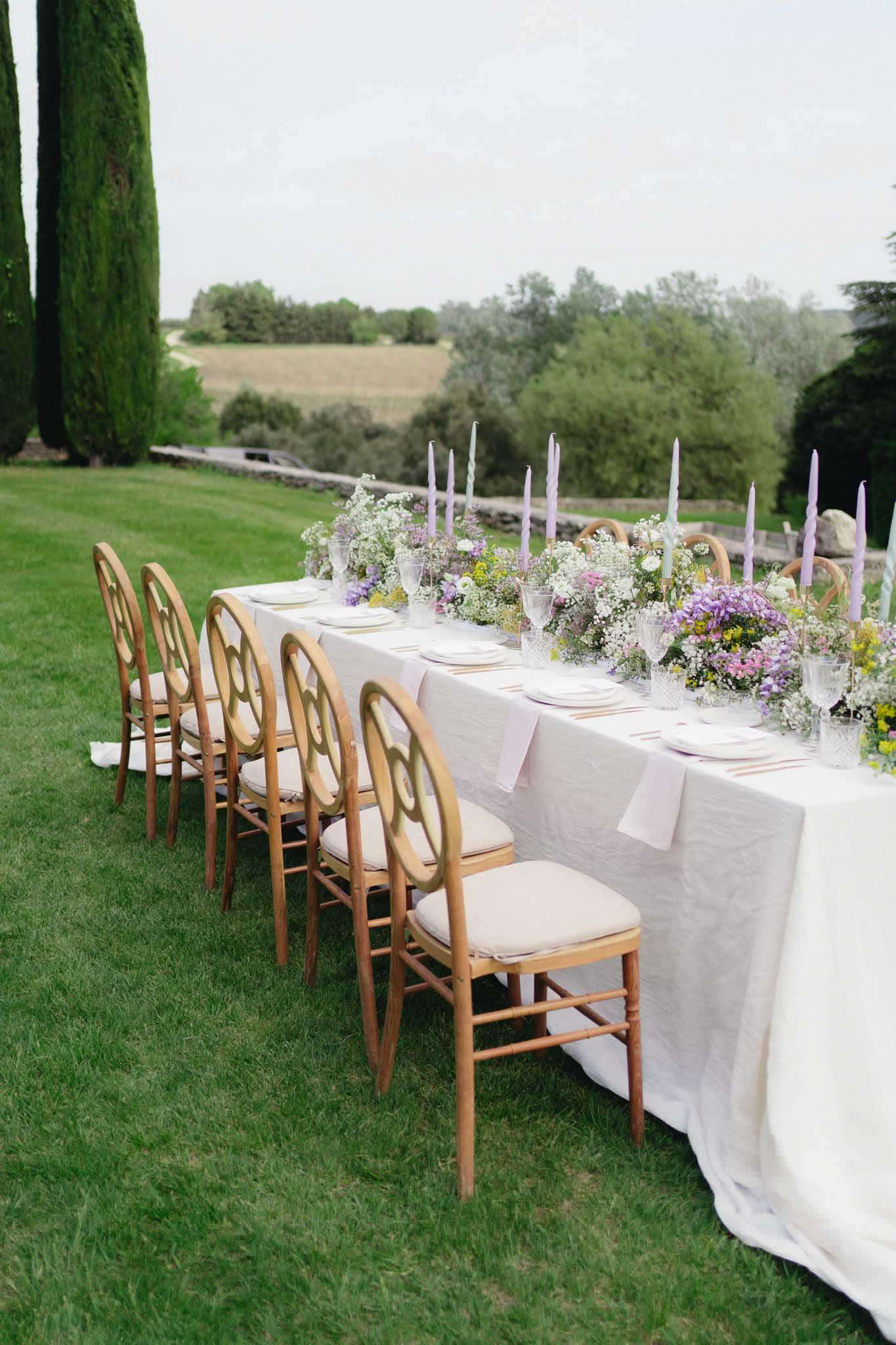Outdoor reception table with white linen, lavender taper candles, and garden-style wildflower centerpiece