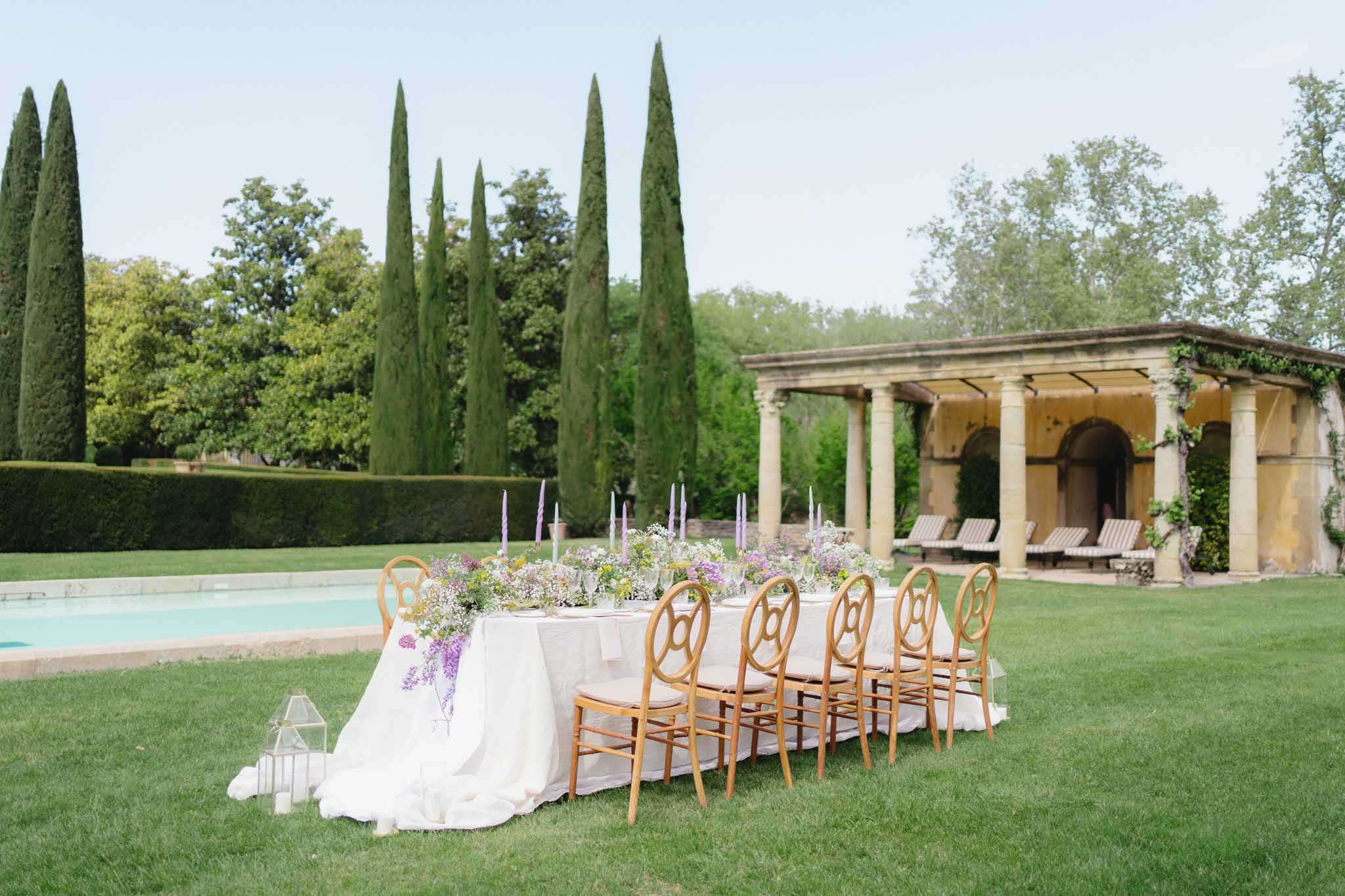 An outdoor reception tablescape set on a lawn beside a rectangular pool, with a stone colonnaded pavilion visible in the background. The long rectangular table is dressed in a white linen cloth that drapes to the ground, topped with a lush runner of lavender, white baby's breath, and small wildflowers, accented by tall lavender-colored taper candles in gold holders. Golden round-back chairs with circular decorative detailing line one side of the table, and small glass lanterns with white pillar candles are placed on the ground near the table ends. The setting has a classic French Provençal aesthetic, with the honey-toned stone pavilion featuring arched openings and columns overgrown with greenery. Wide shot capturing the full table arrangement and surrounding estate grounds. Potential venue feature image.