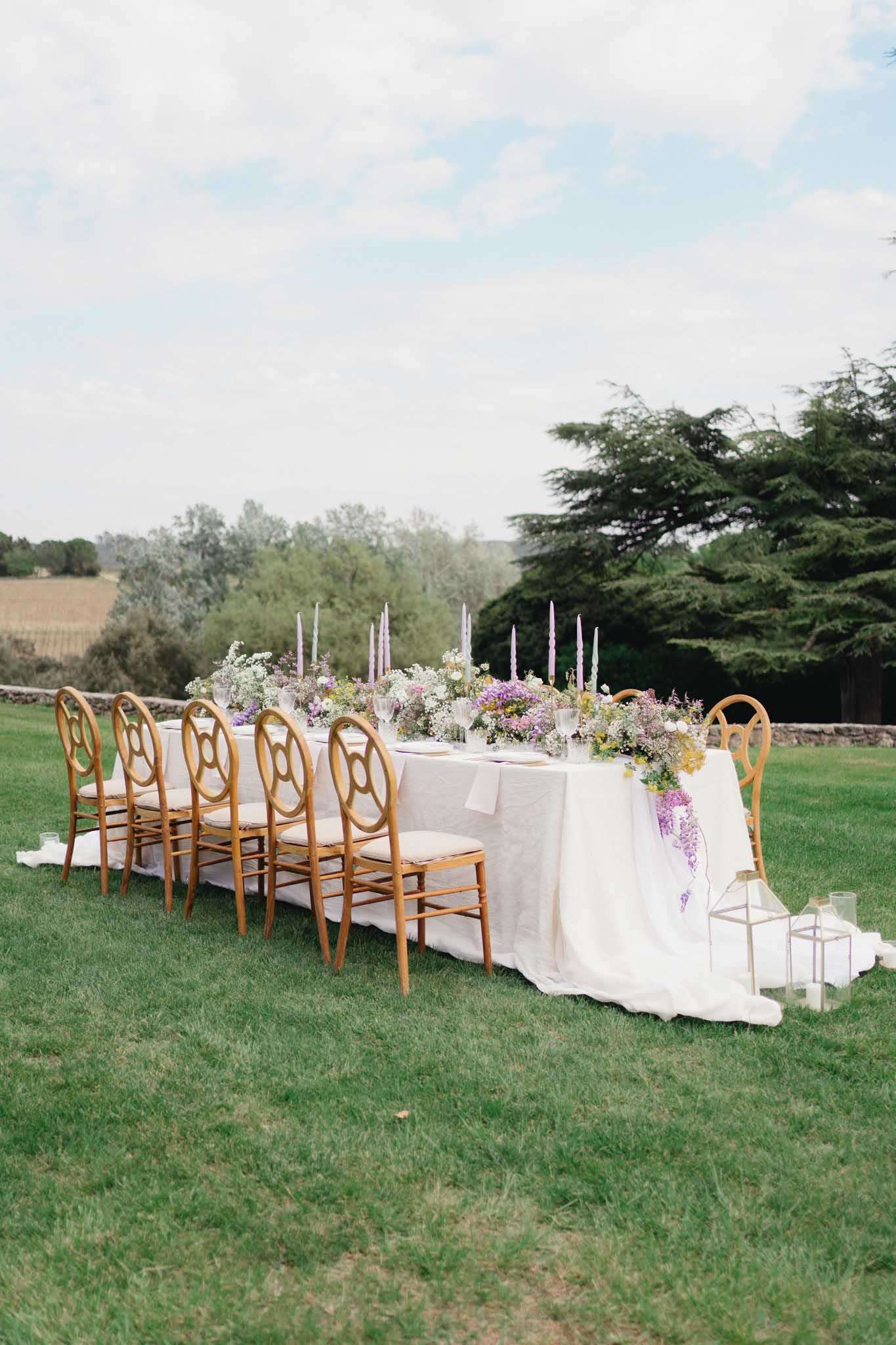 Outdoor reception table on lawn with garden-style floral runner of wisteria and wildflowers, gold chairs, and lilac taper ...